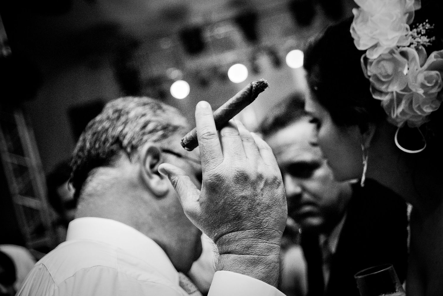 Black and white candid photo of a man smoking a cigar at a wedding reception event.