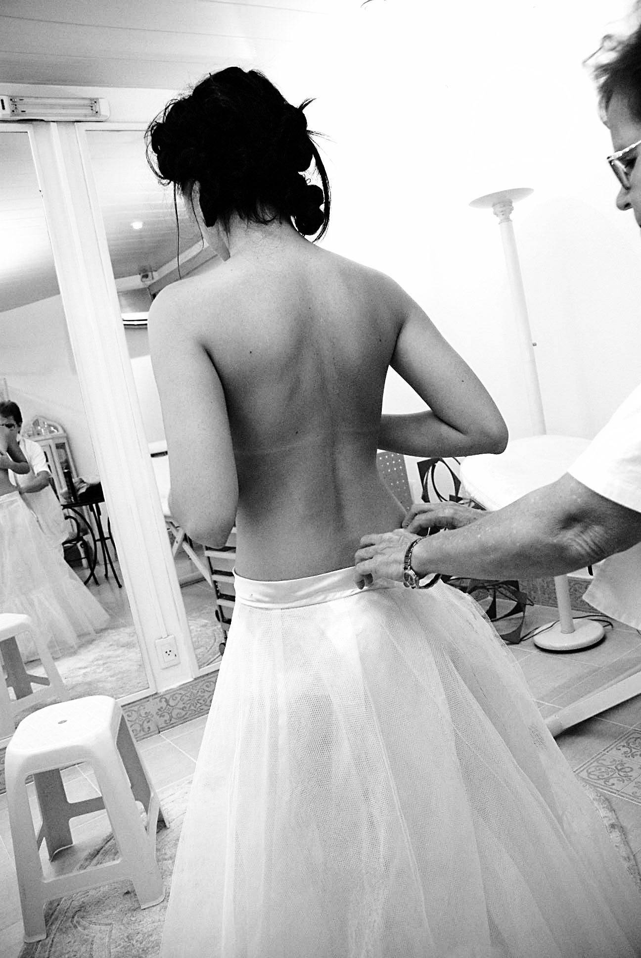 Black and white photo of a bride having her backless wedding gown fastened by a helper.