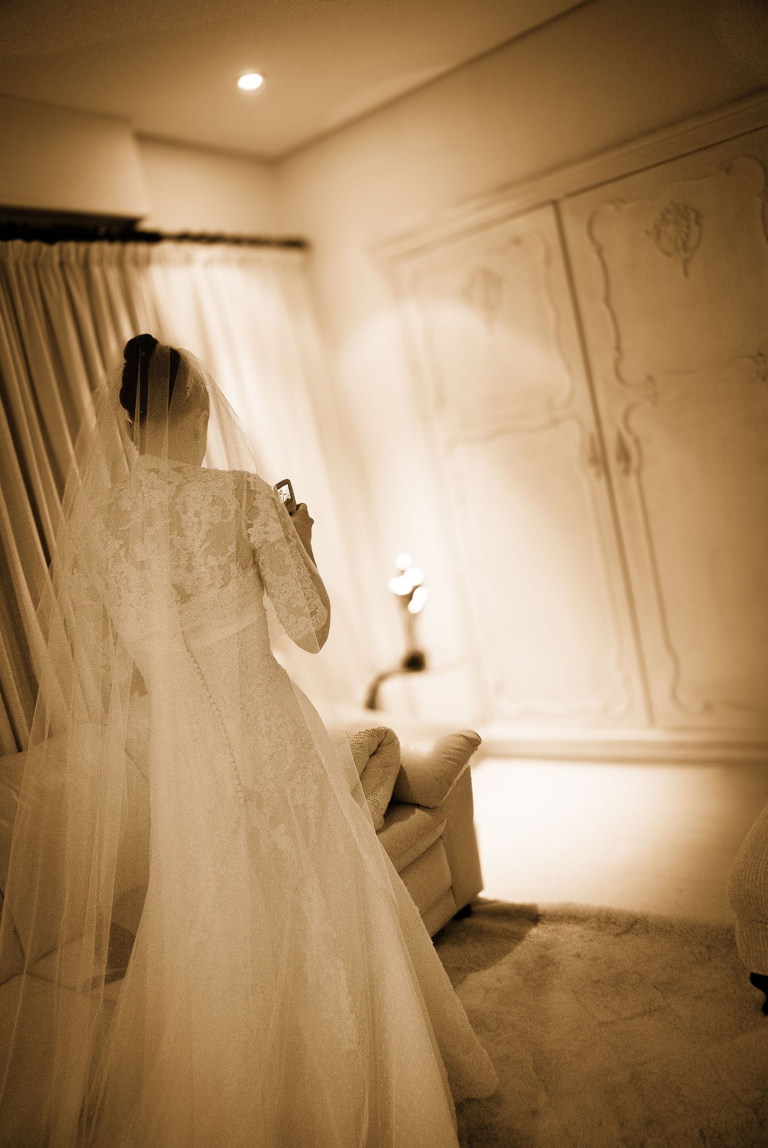 Bride in lace wedding gown with veil standing in warmly lit elegant room before ceremony.