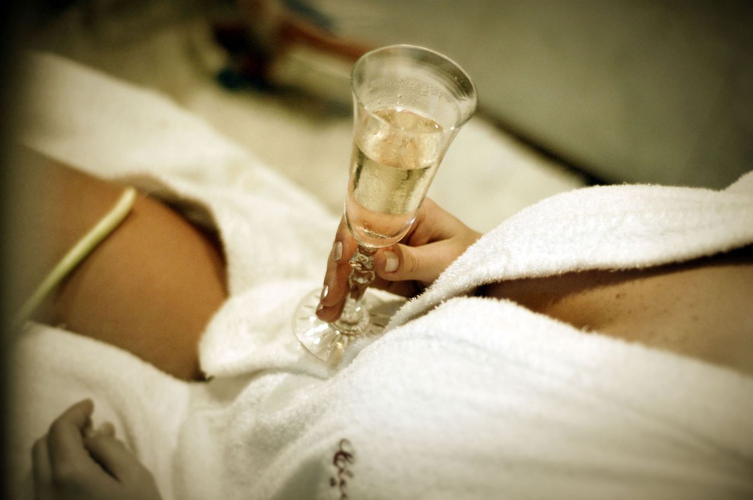 Bride in white robe holding champagne flute while relaxing during wedding preparations.