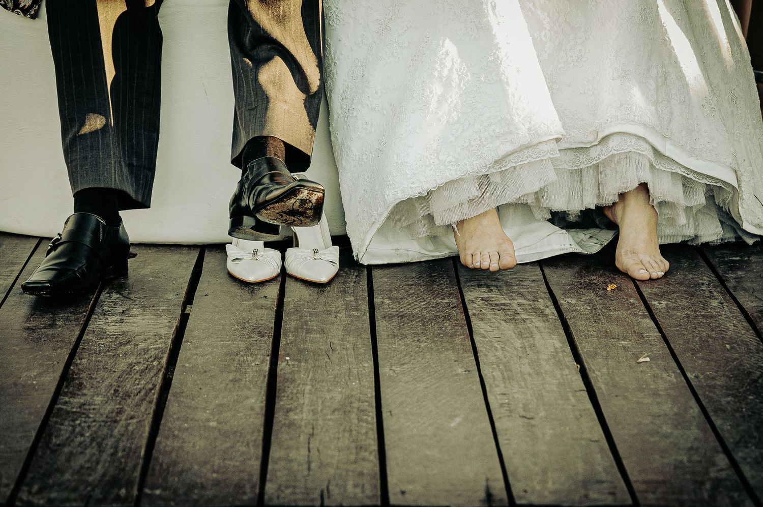 Close-up of wedding party feet on wooden deck with bride barefoot beside groom in formal shoes.