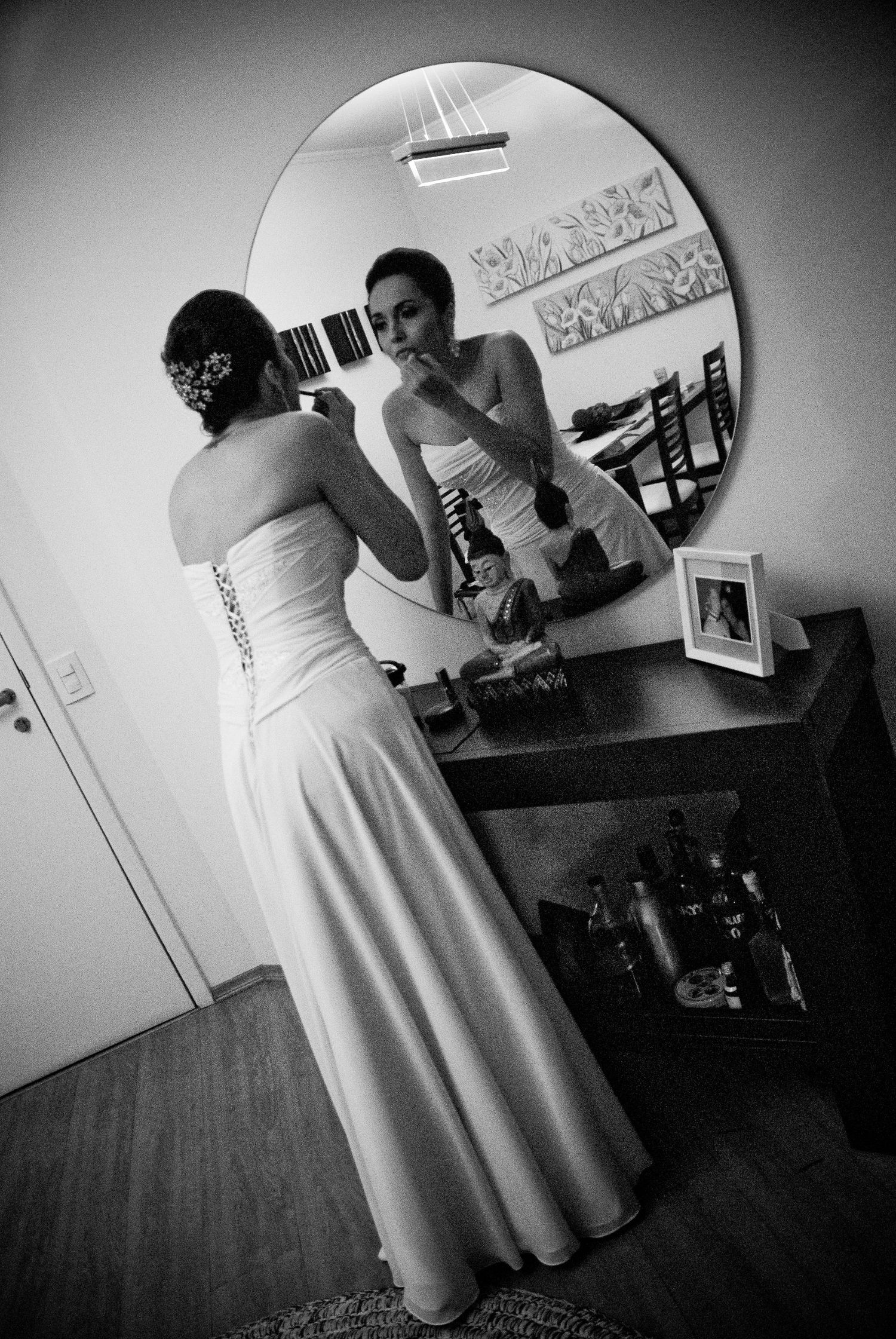 Bride in strapless white gown applying makeup at vanity mirror in black and white photo.