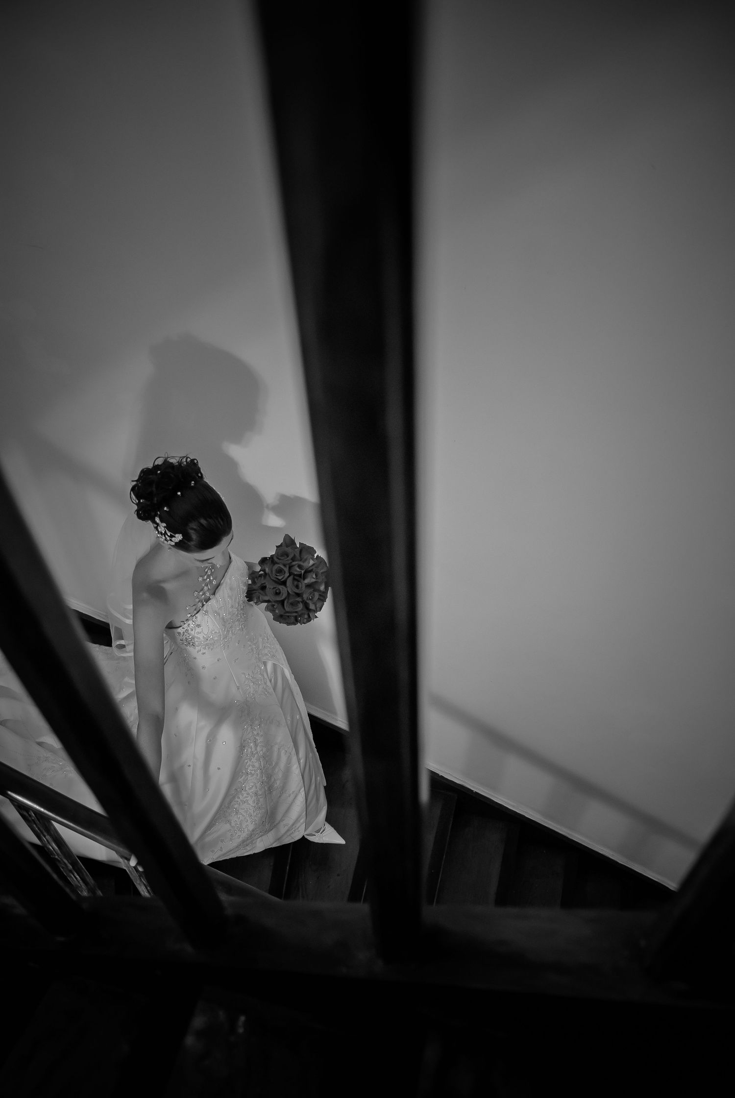 Bride in white gown holding bouquet descending staircase captured in dramatic black and white.