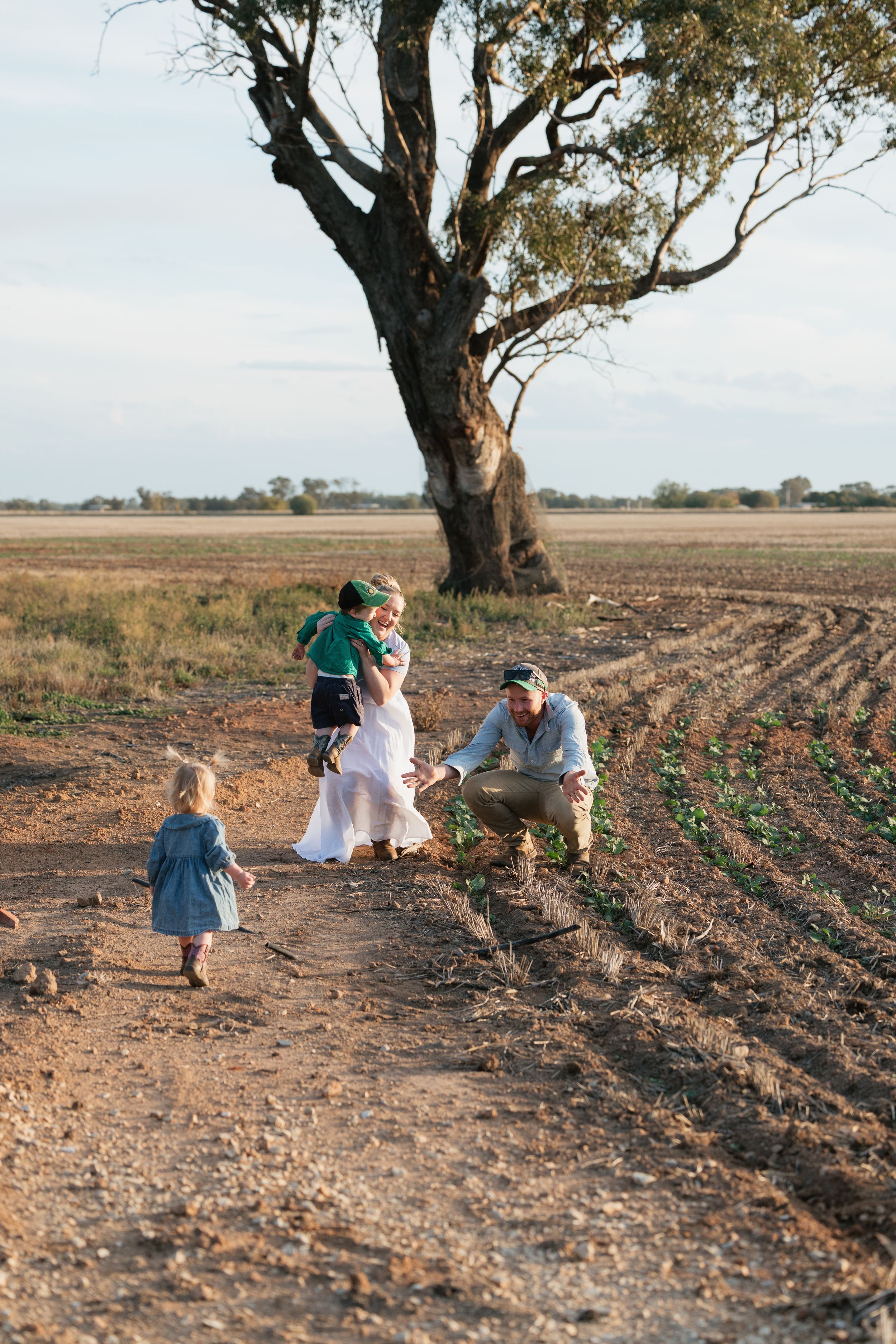 Family with young children exploring a rural farm field beside a large gum tree in the Australian outback.