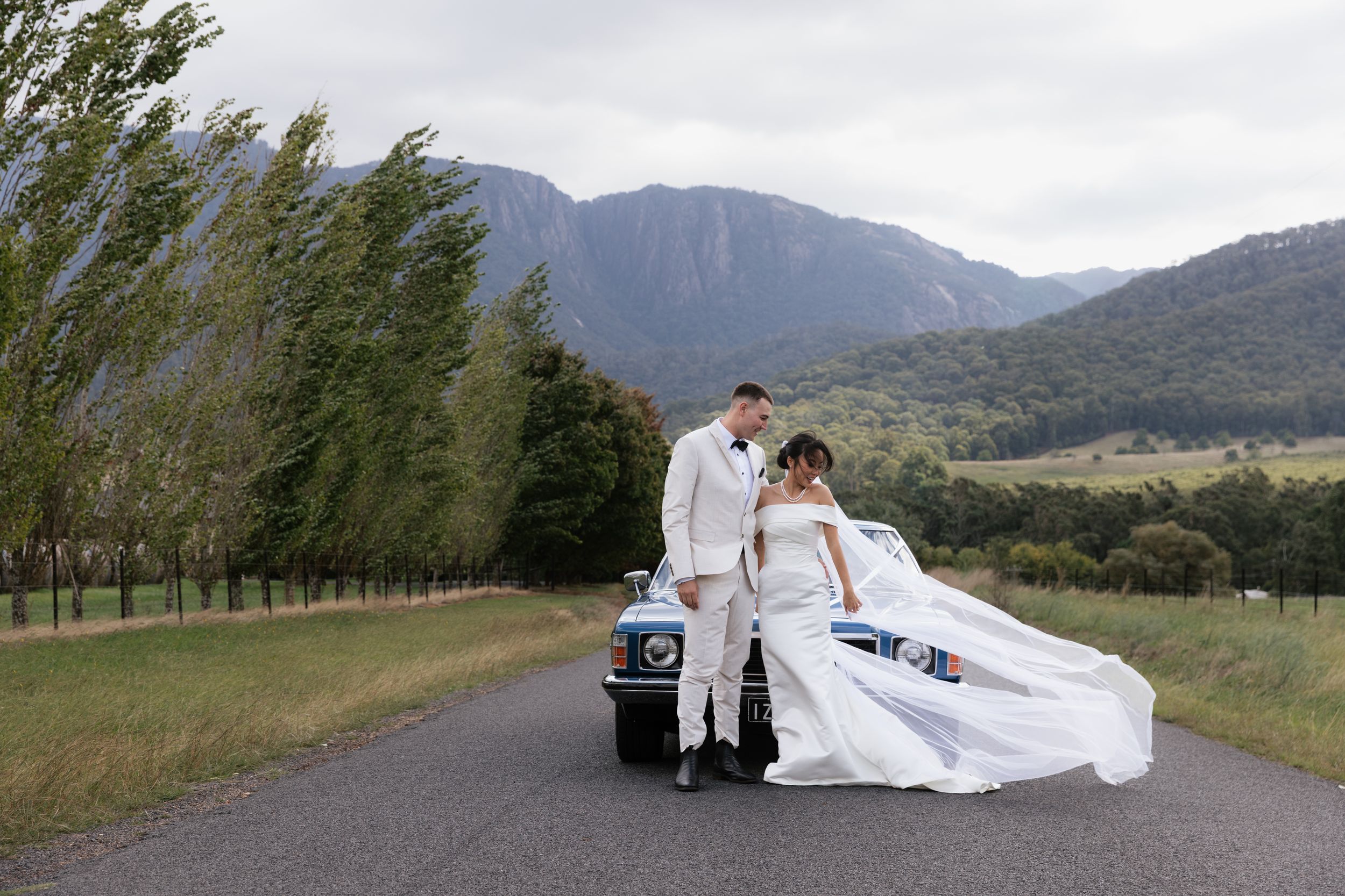 Bride and groom pose beside a vintage blue car on a scenic mountain road with lush green hills and trees in the background.