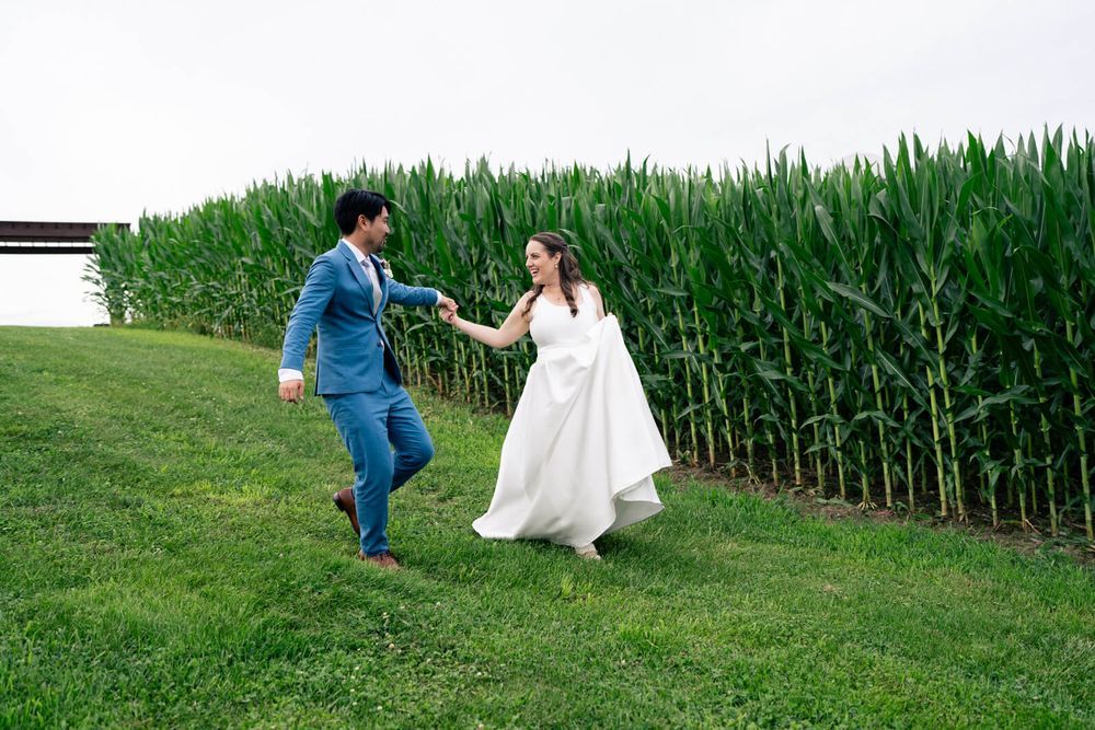 Bride and groom holding hands running through green grass beside tall cornfield on their wedding day.