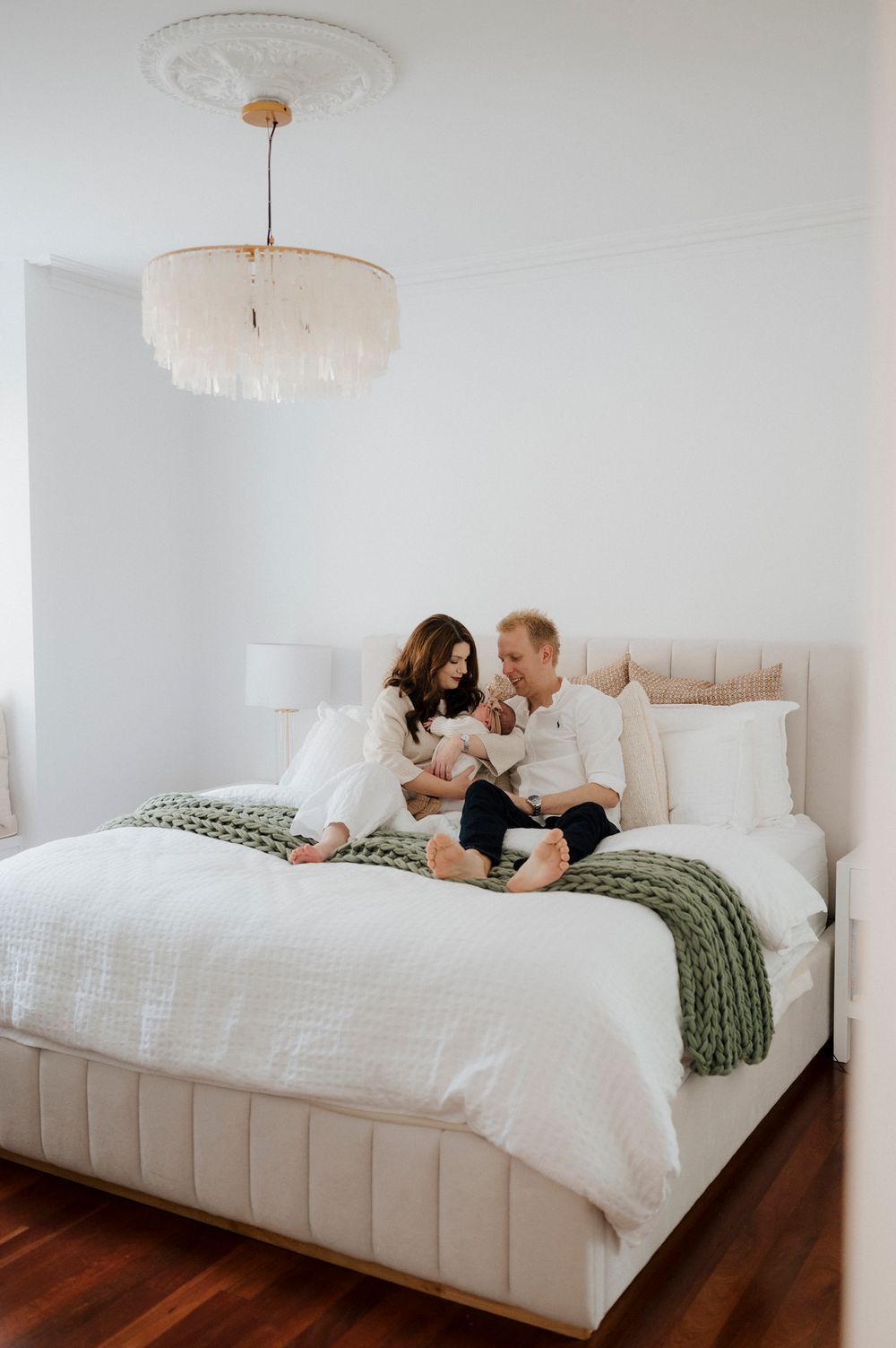New parents sitting on a white bed with green knit blanket, cradling their newborn baby in a bright bedroom.