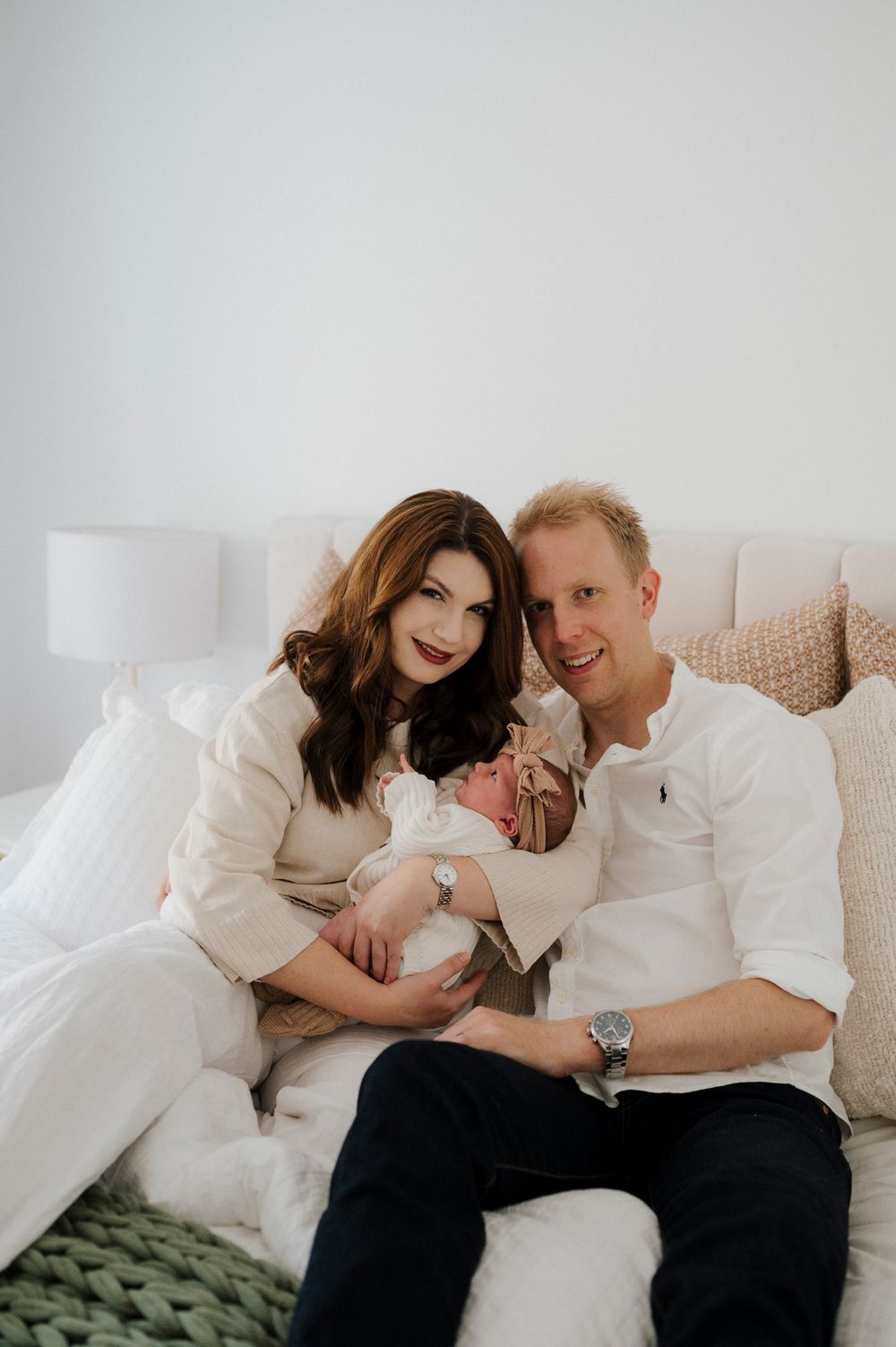 A couple sitting on a white bed smiling and holding their newborn baby, dressed in casual white outfits.