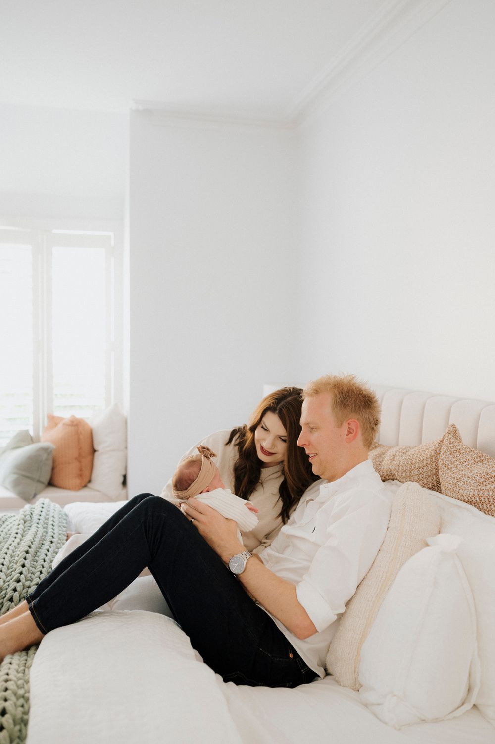 Young couple sitting together on a white bed, lovingly gazing at their newborn baby in a bright room.