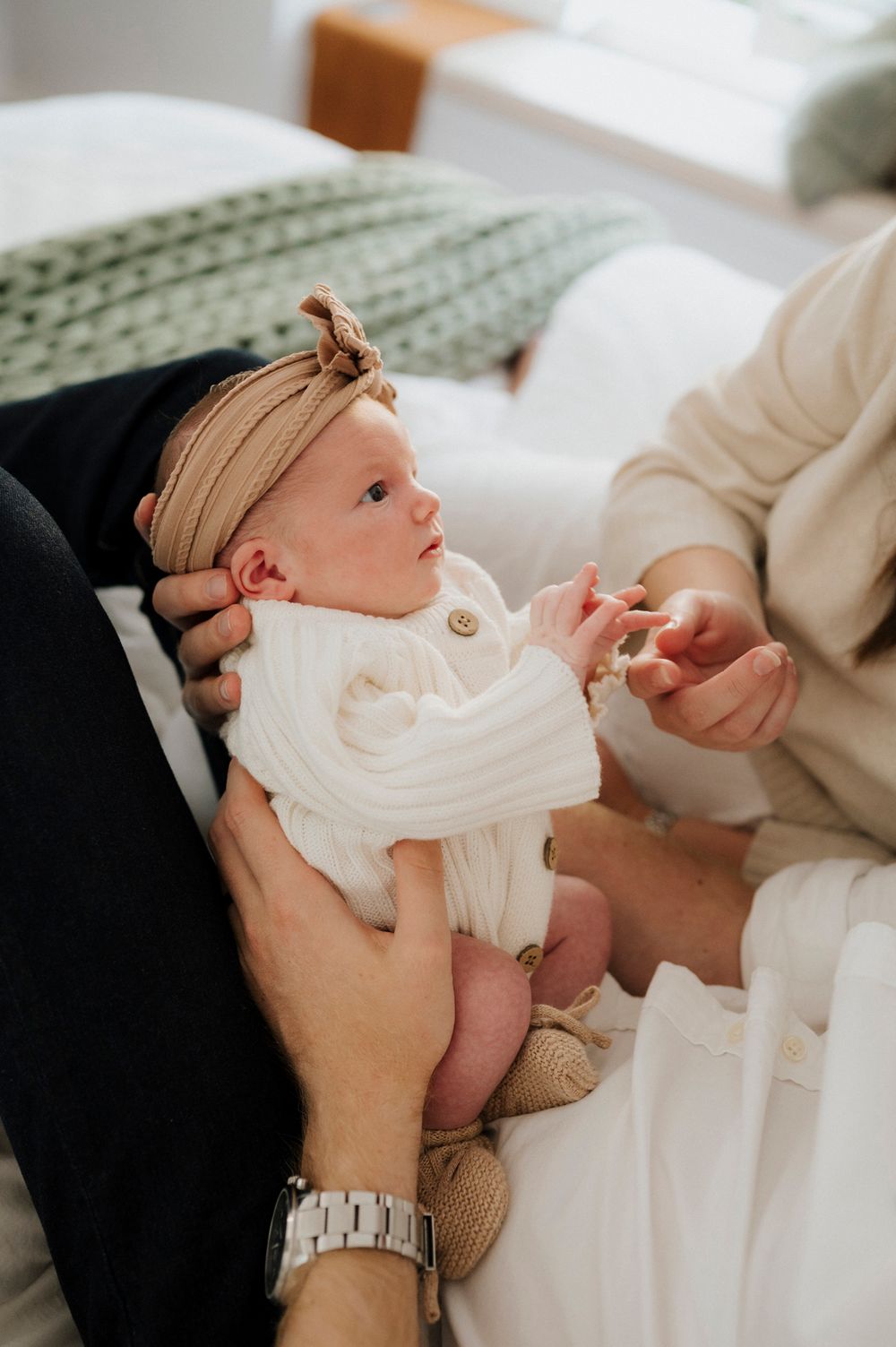 Newborn baby in white knit outfit and tan bow headband being held by parents on a white bed.
