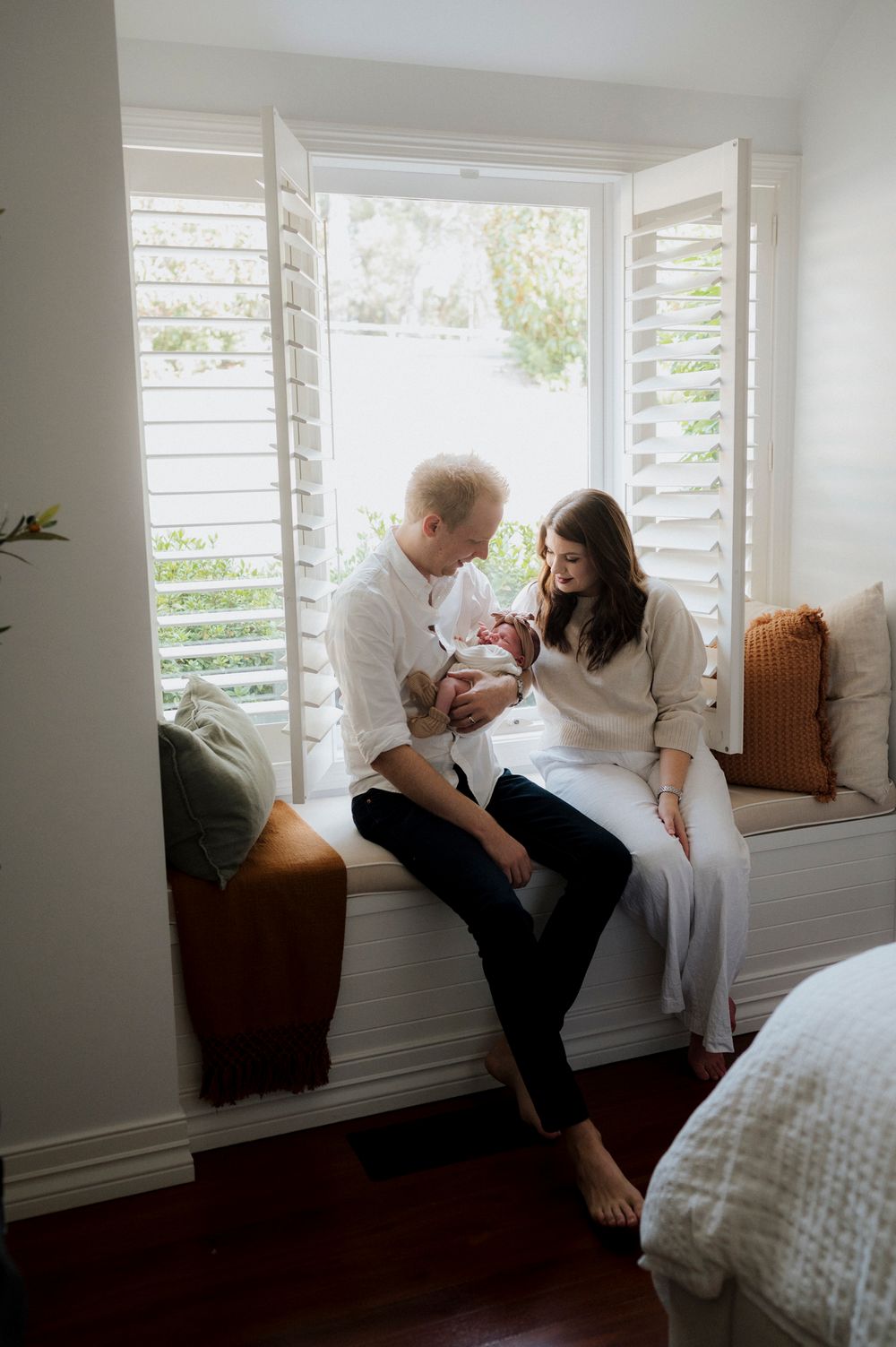 New parents sitting on a window seat, lovingly gazing at their newborn baby in a bright, cozy bedroom.