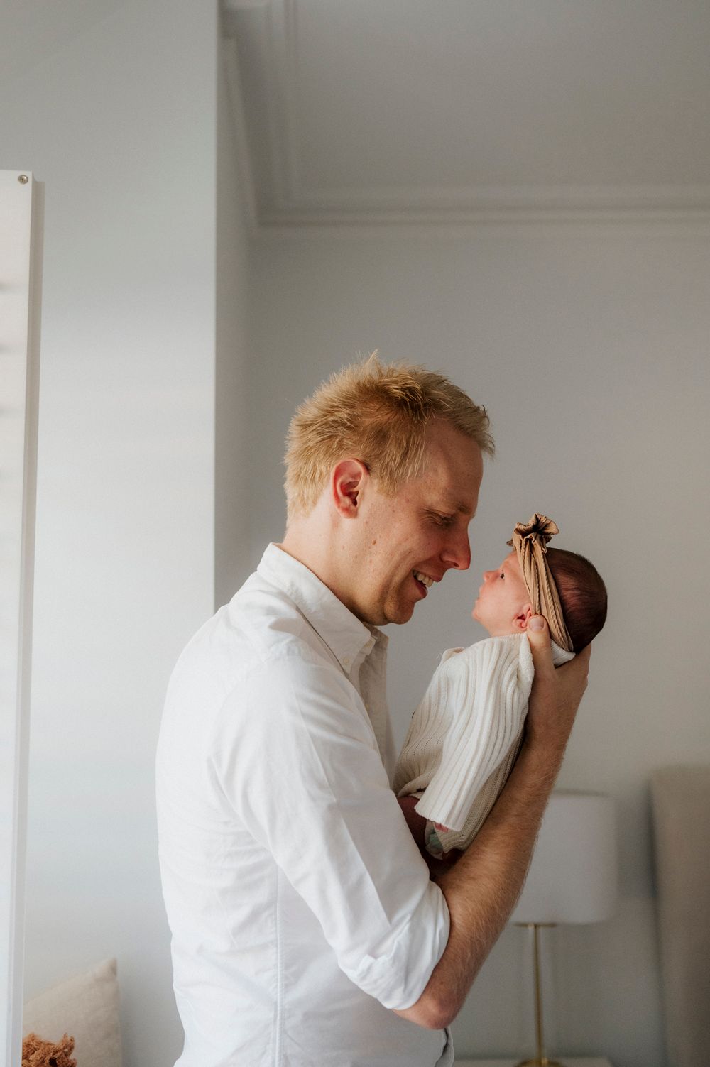 A smiling father in a white shirt tenderly holds his newborn baby face-to-face in a bright, softly lit room.