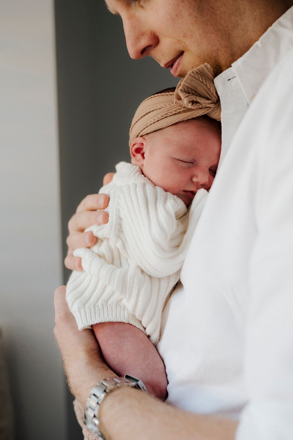 Parent tenderly holding a sleeping newborn baby dressed in white knit outfit with tan bow headband.