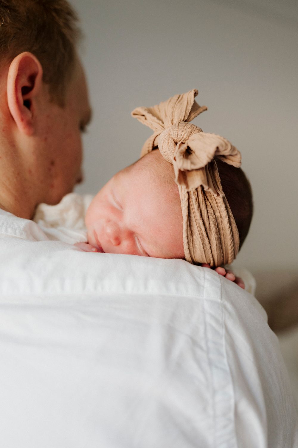 A newborn baby with a tan bow headband resting on an adult's shoulder in a tender moment.