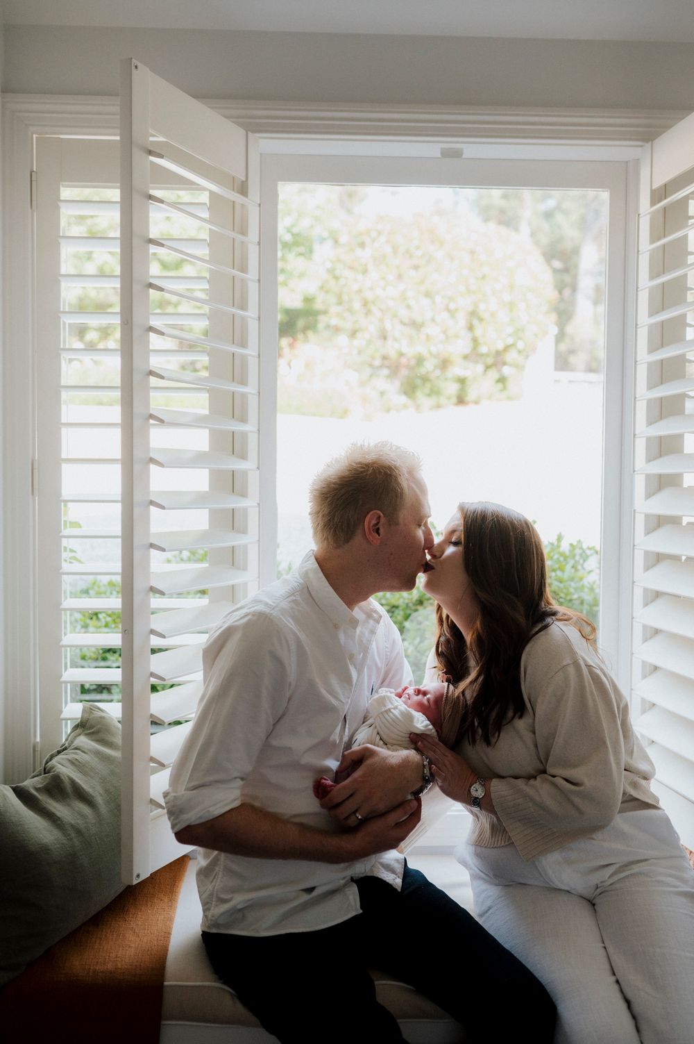 Couple sharing a tender kiss while holding their newborn baby by a bright window with white plantation shutters.