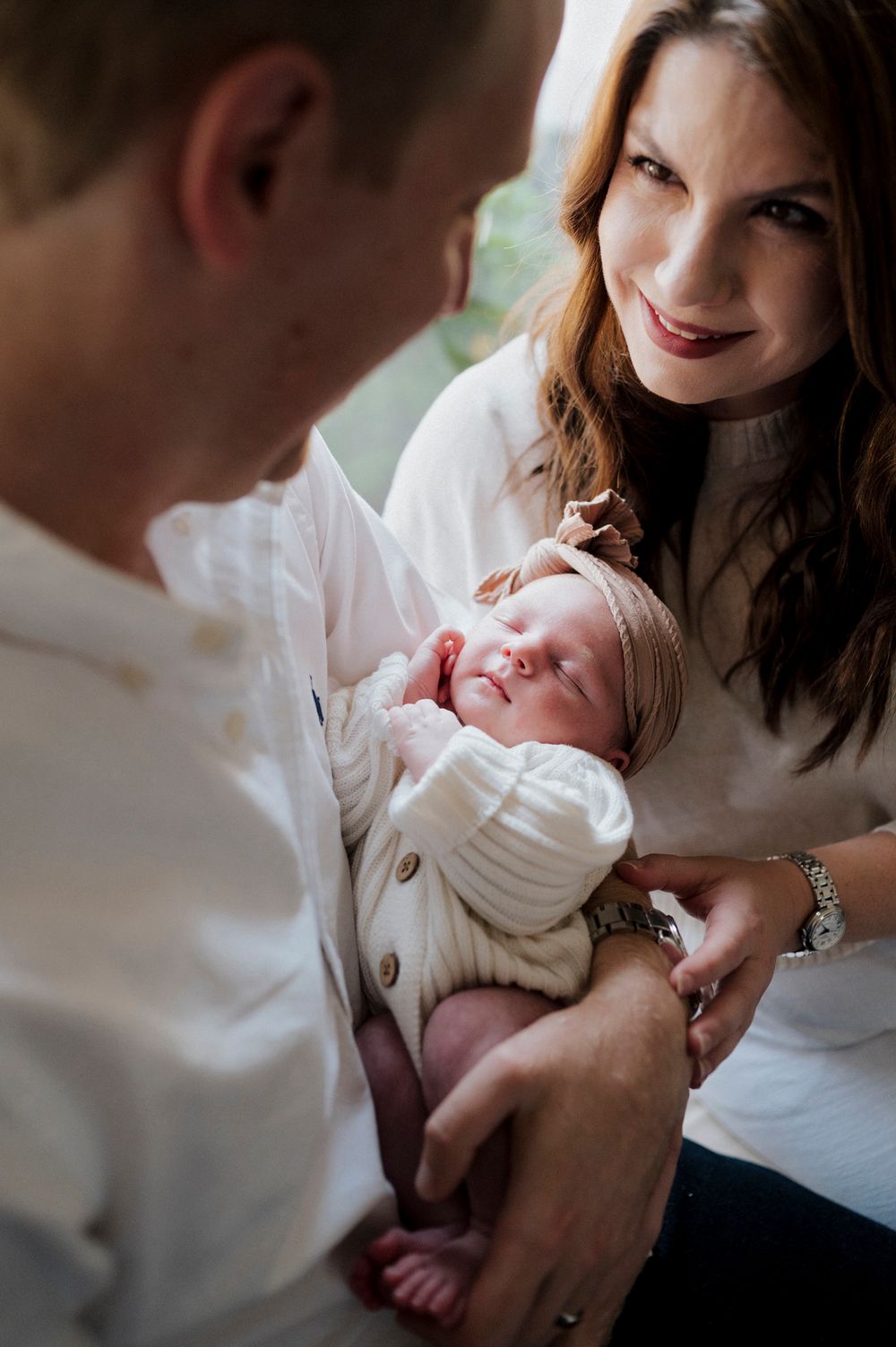 Parents in white clothing lovingly gaze at their sleeping newborn baby during an intimate family portrait session.