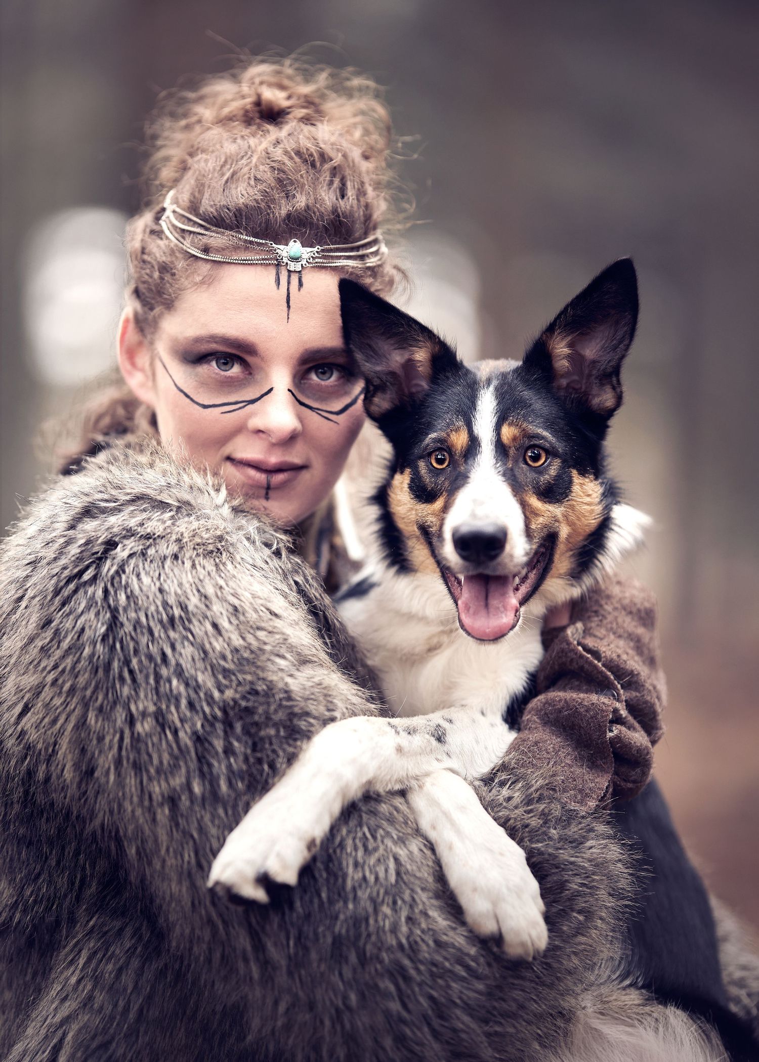 Viking woman with face paint and crown holds a tricolor Border Collie dog in a forest setting.