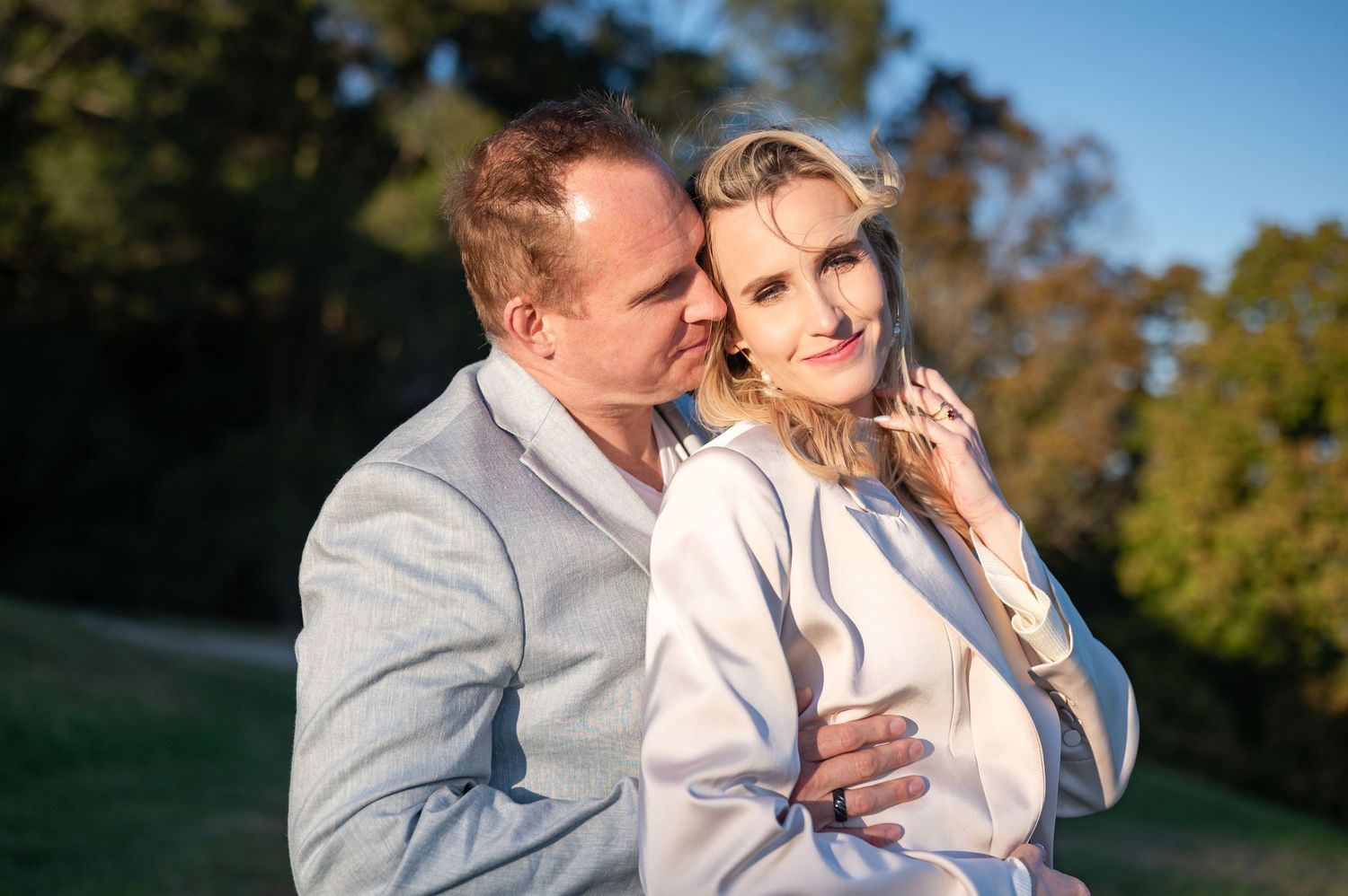 A couple embraces outdoors, woman in white top touching her necklace, man in grey jacket, surrounded by green trees.