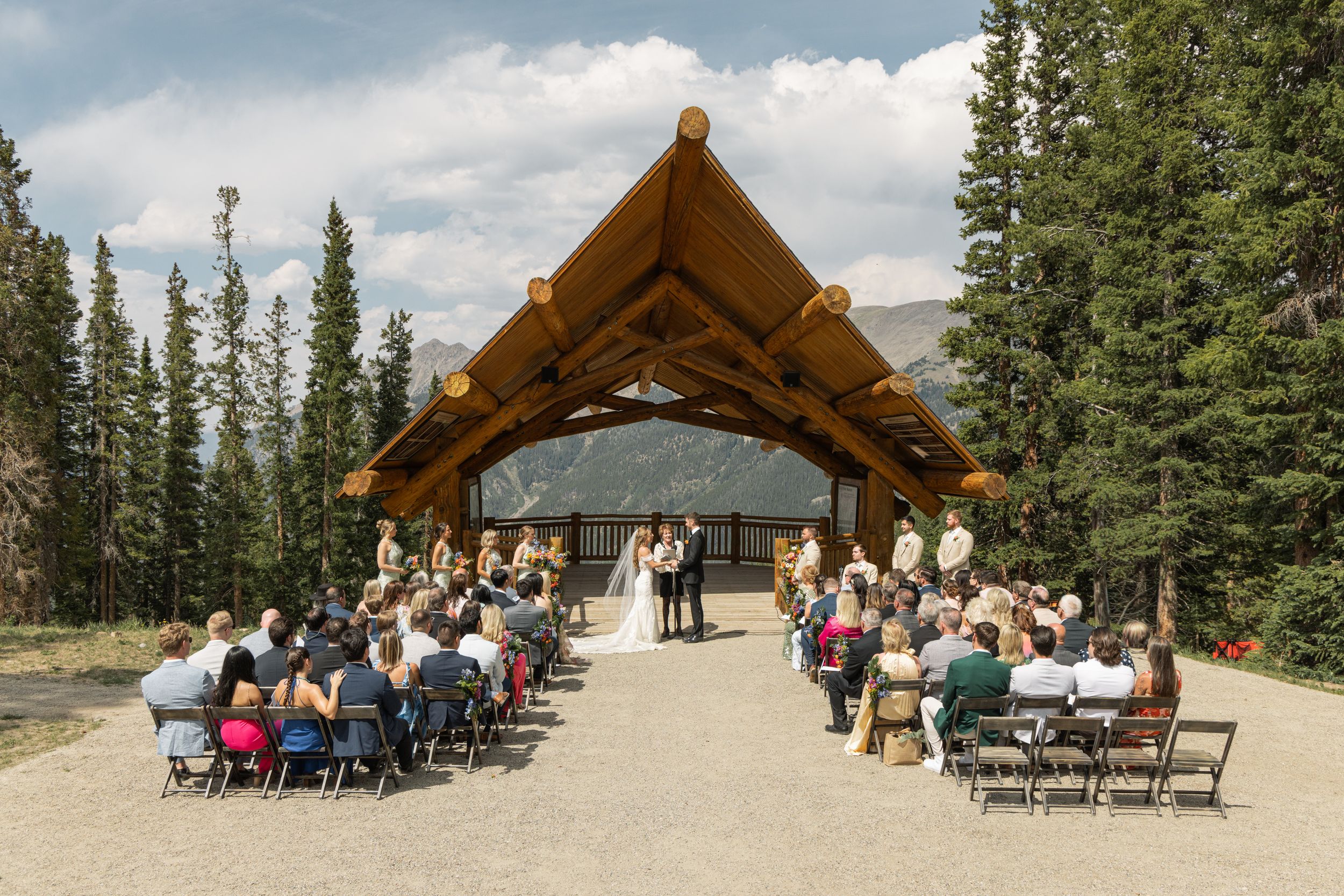 Outdoor wedding ceremony under a rustic log timber frame pavilion surrounded by pine trees and mountains.
