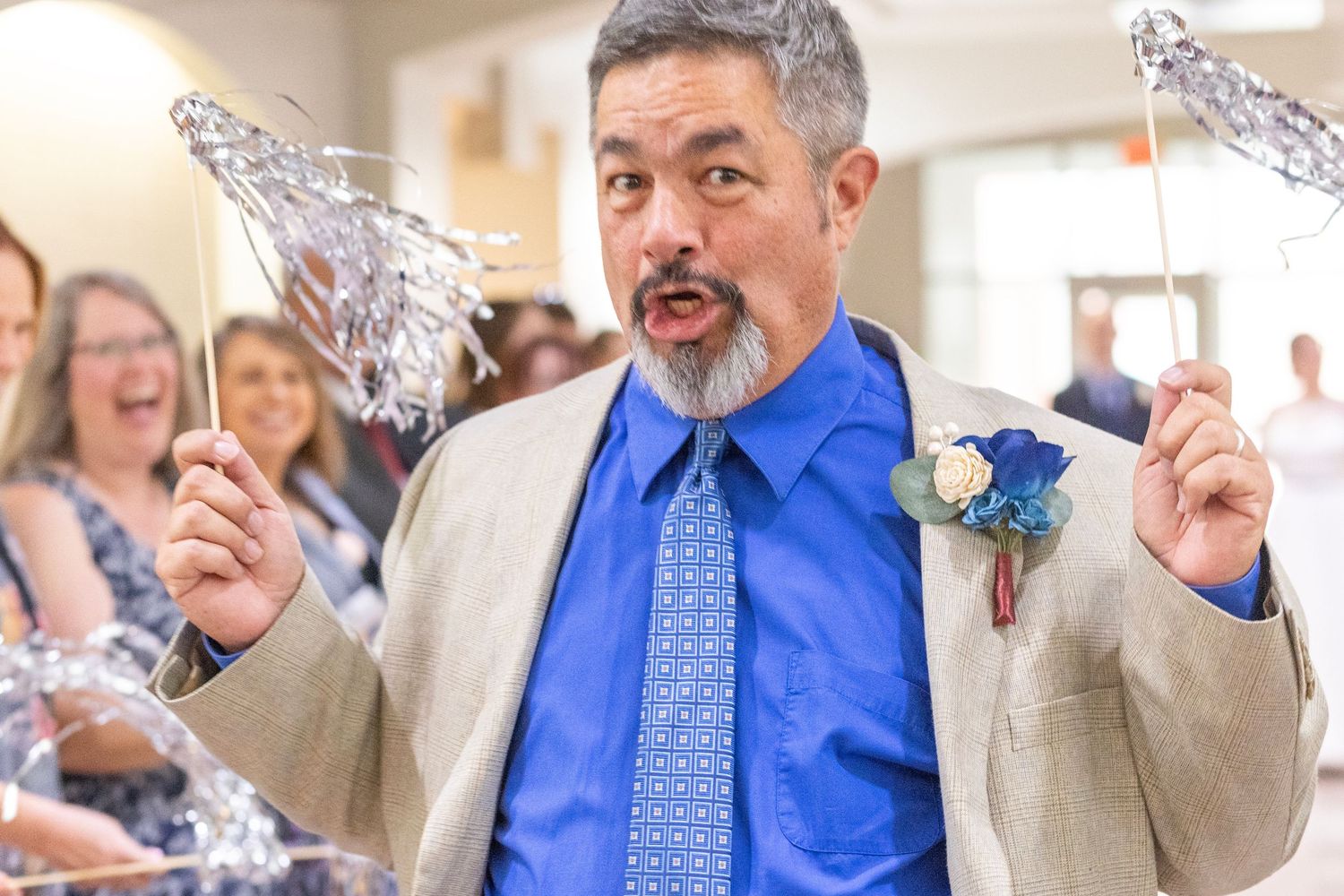 Man in blue shirt and tan suit holds decorative wire flowers at a wedding celebration.