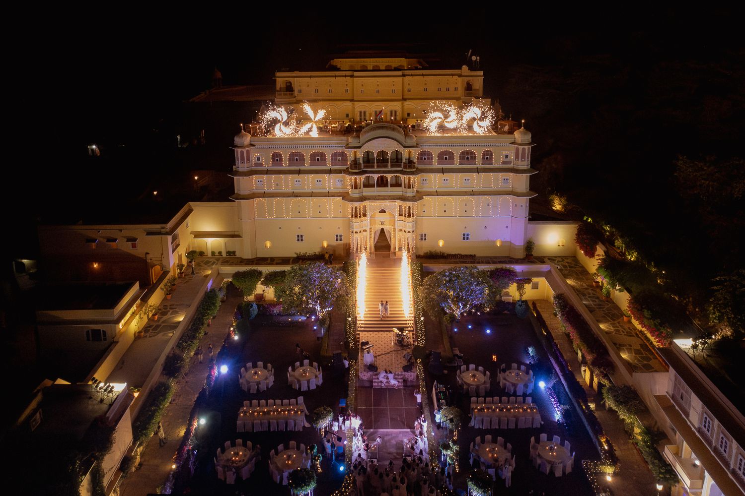 Aerial night view of an illuminated Indian palace with ornate architecture, glowing lights, and an elegant outdoor event setup.