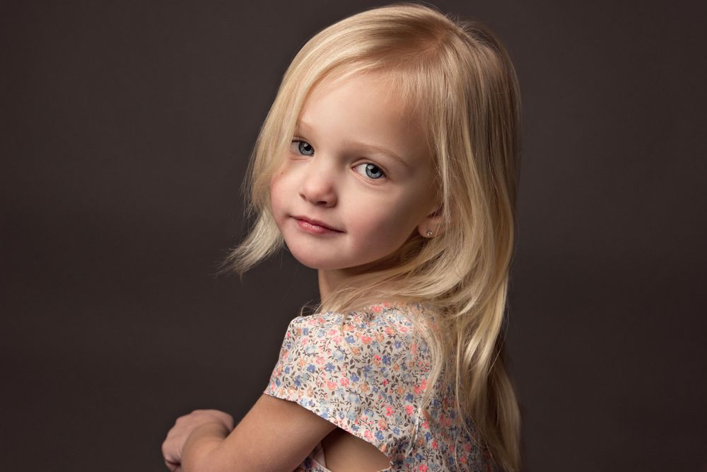 Adorable young blonde girl in a floral dress posing with a gentle smile against a dark studio background.