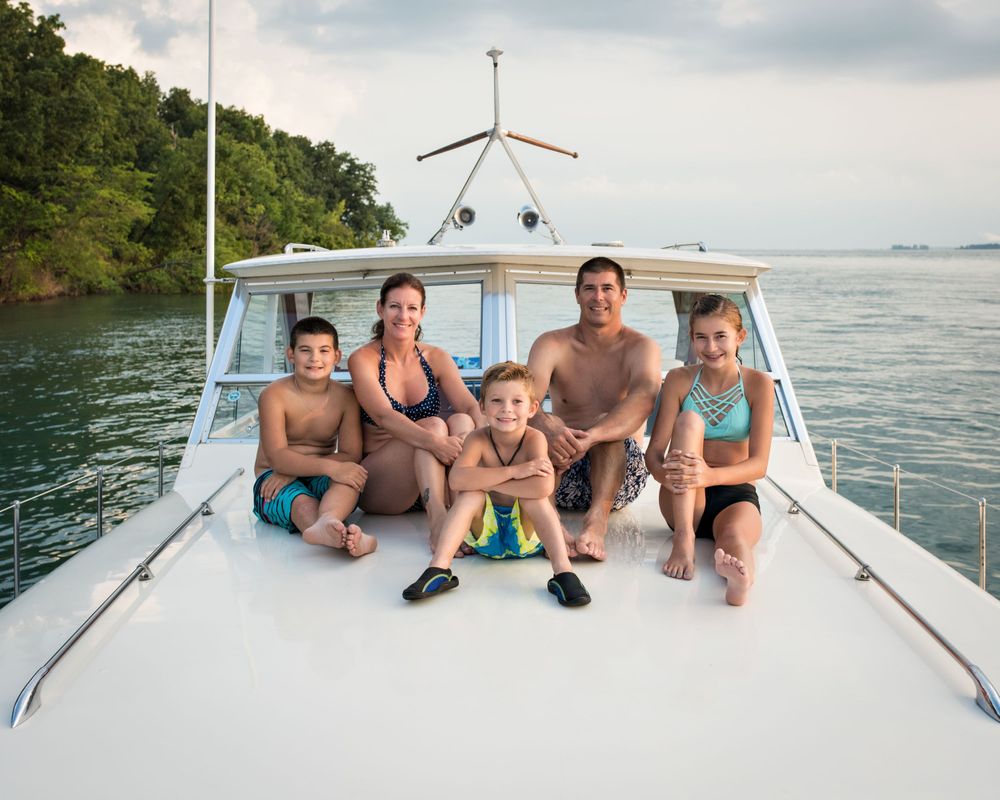 Family of five in swimwear sitting together on the bow of a boat surrounded by calm green waters.