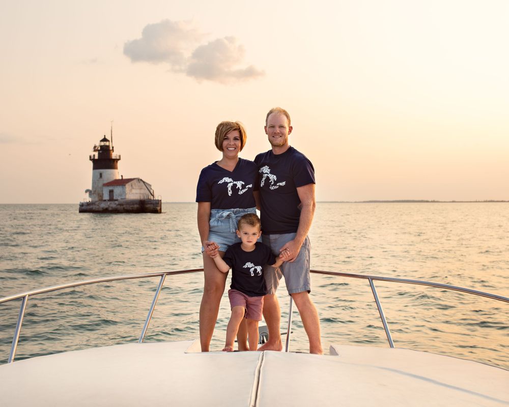 Happy family of three in matching navy shirts posing on a boat bow with a lighthouse at sunset.