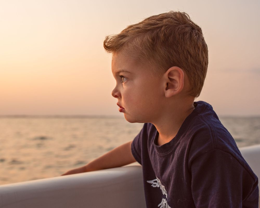 Young boy in a navy shirt gazing thoughtfully at the ocean horizon during a beautiful golden sunset.