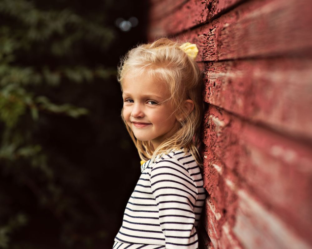 Smiling blonde girl leaning against a red wooden wall outdoors.
