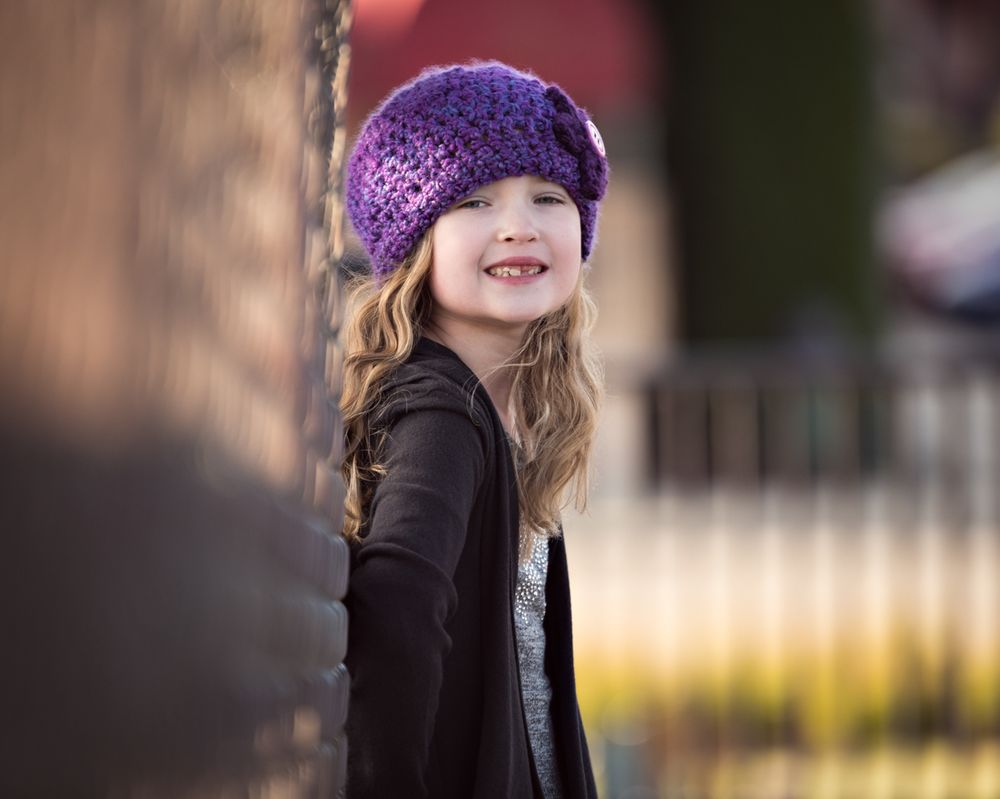 Happy girl in purple crochet hat smiling beside a brick wall.