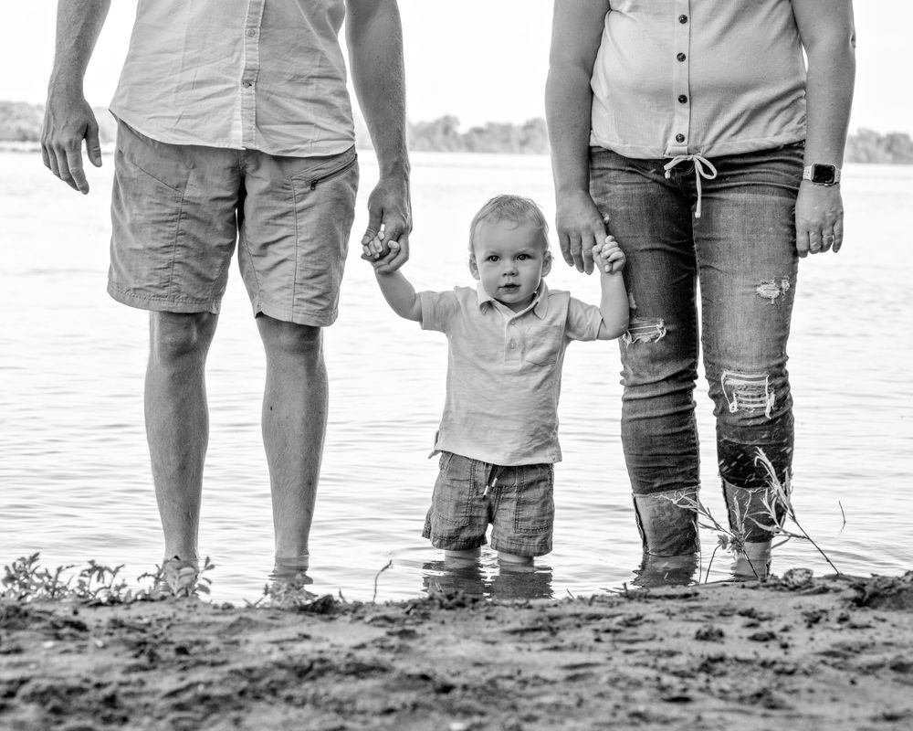 Black and white photo of parents holding hands with their toddler standing between them at a sandy shoreline.