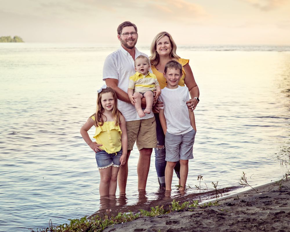 Family of five standing at the water's edge at sunset, smiling together in coordinating yellow and white outfits.