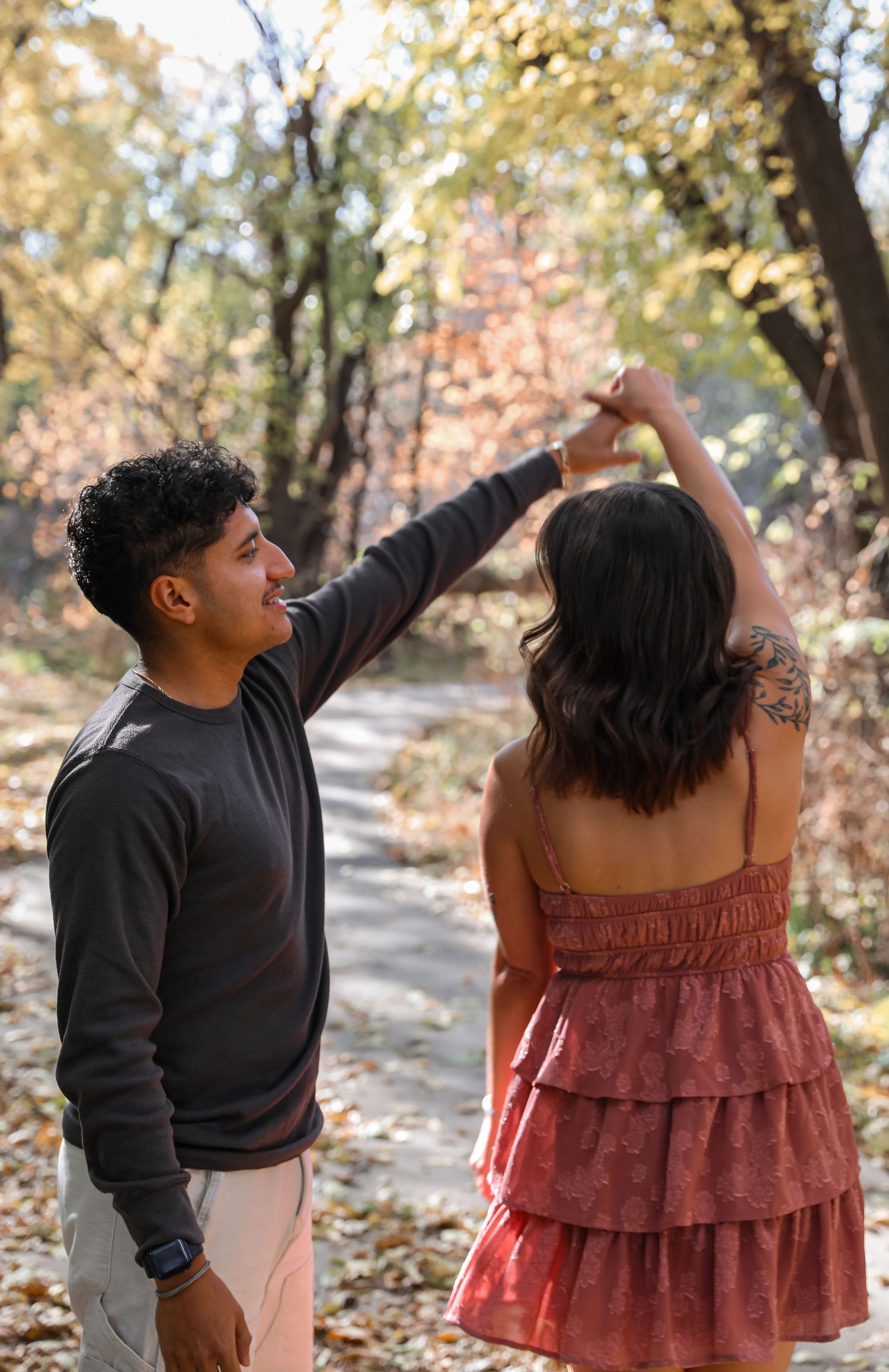 A couple dances together on a forest path surrounded by autumn foliage, the man spinning the woman in a red dress.