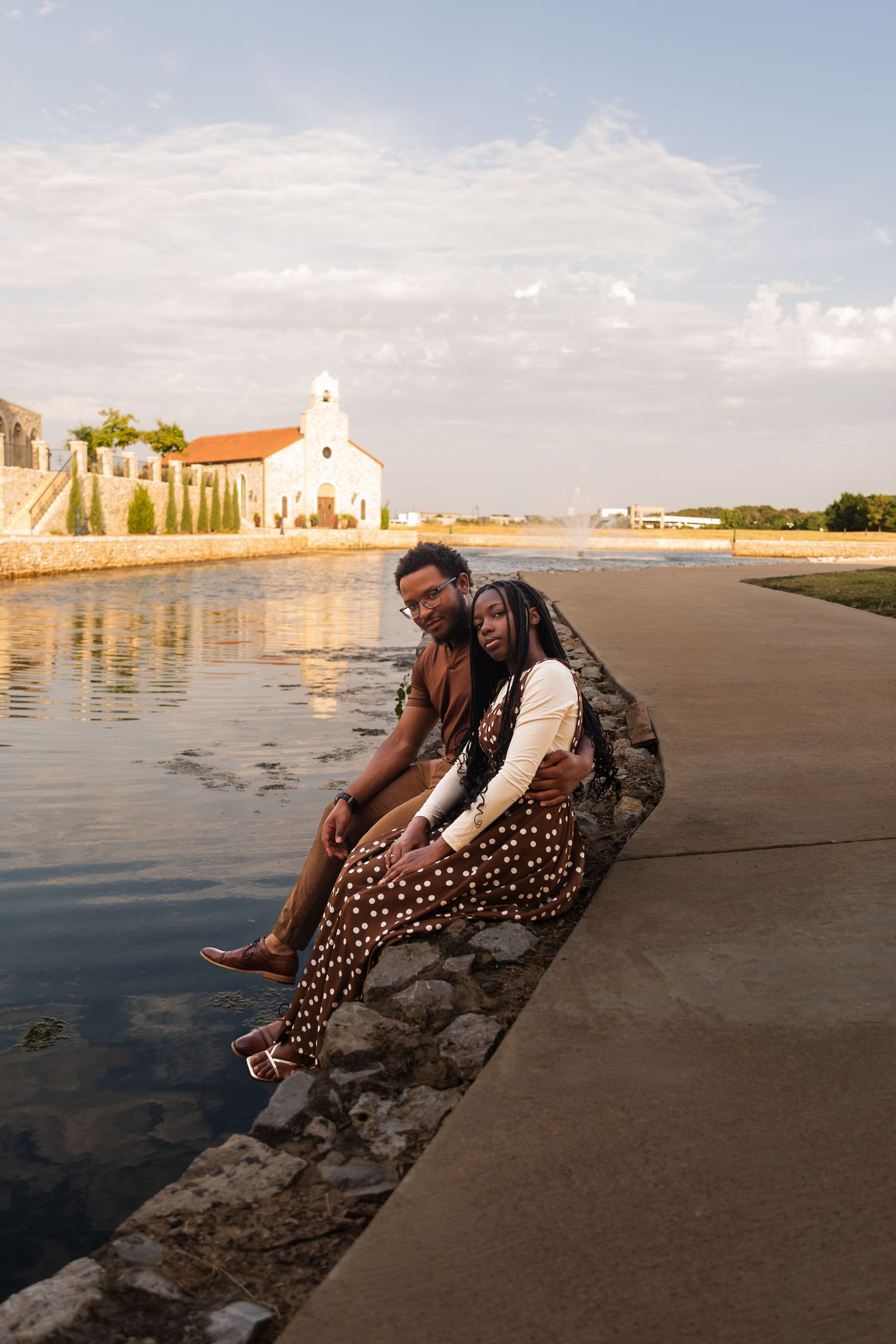A couple sits together on a stone ledge by a calm waterway, with a white church and golden sunset in the background.