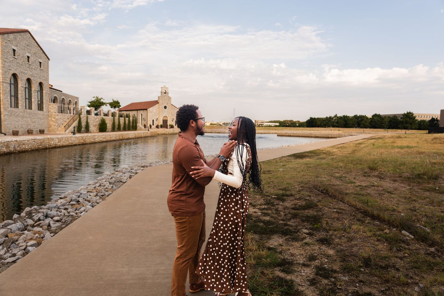 Couple embracing by a scenic waterway near a historic building on a cloudy day outdoors.