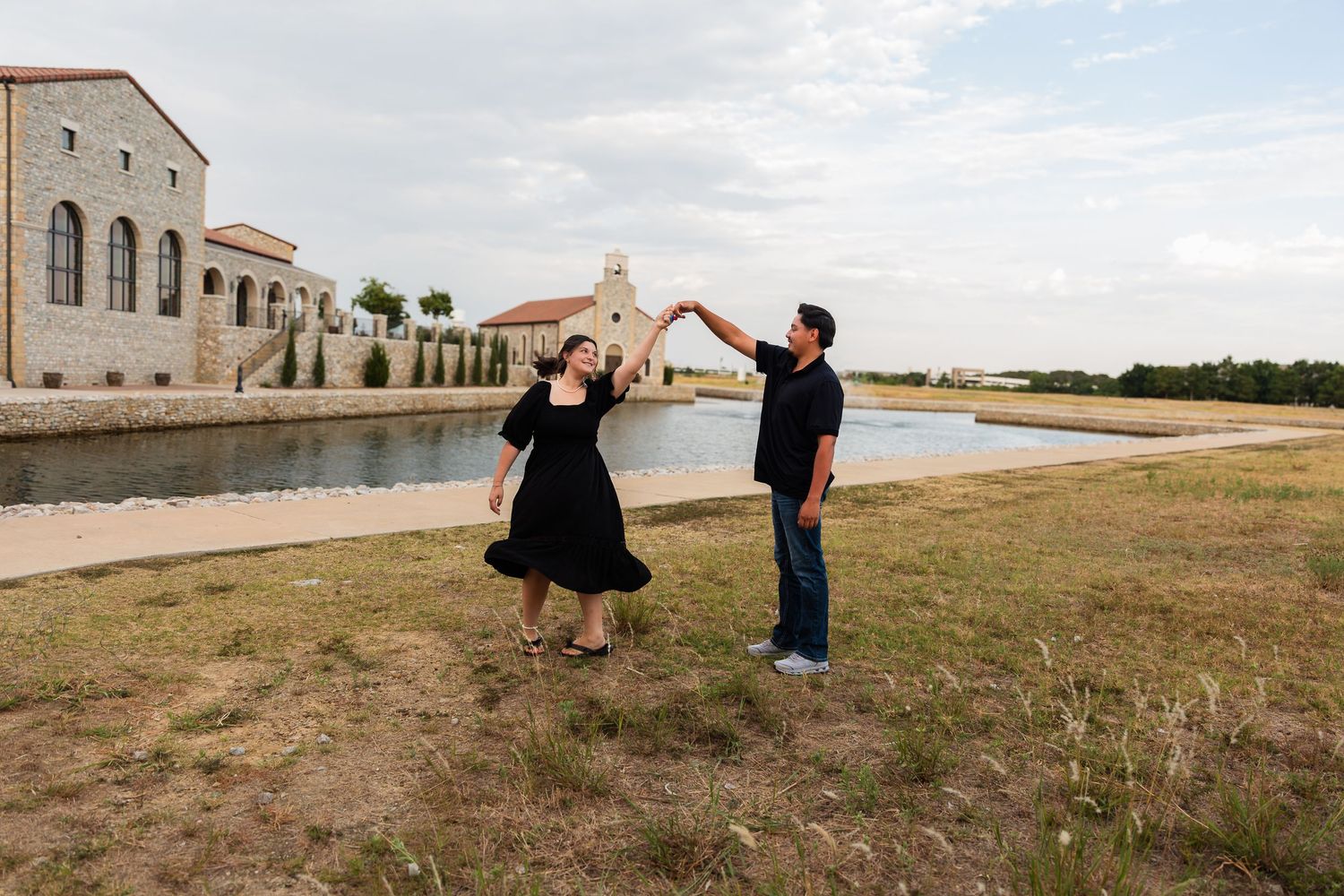 Couple dancing outdoors near a stone building and reflective pond, woman in black dress being twirled by man in casual attire.