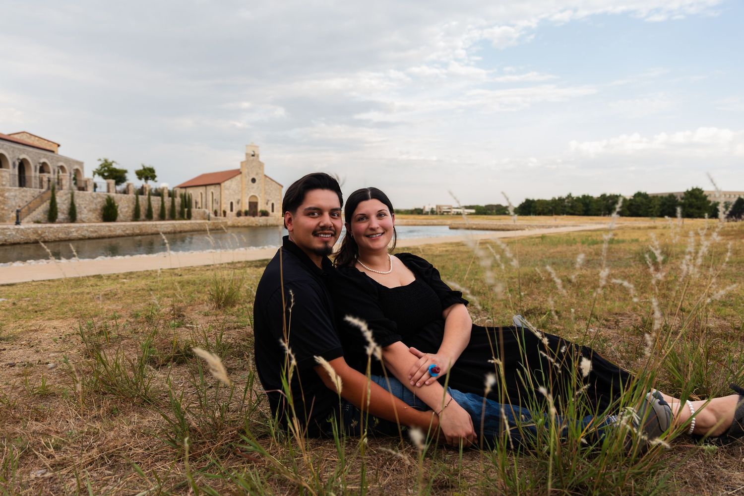 Couple sitting together in grass near a scenic pond and historic building under a cloudy sky.