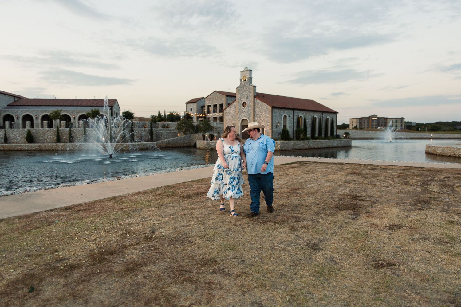 Couple posing in front of a scenic pond with fountains and a historic building at dusk in Texas.