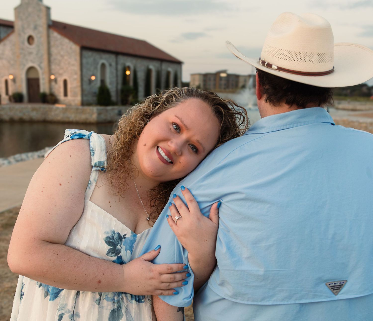 A smiling woman in a floral dress hugs a man in a cowboy hat outdoors near a stone building at sunset.