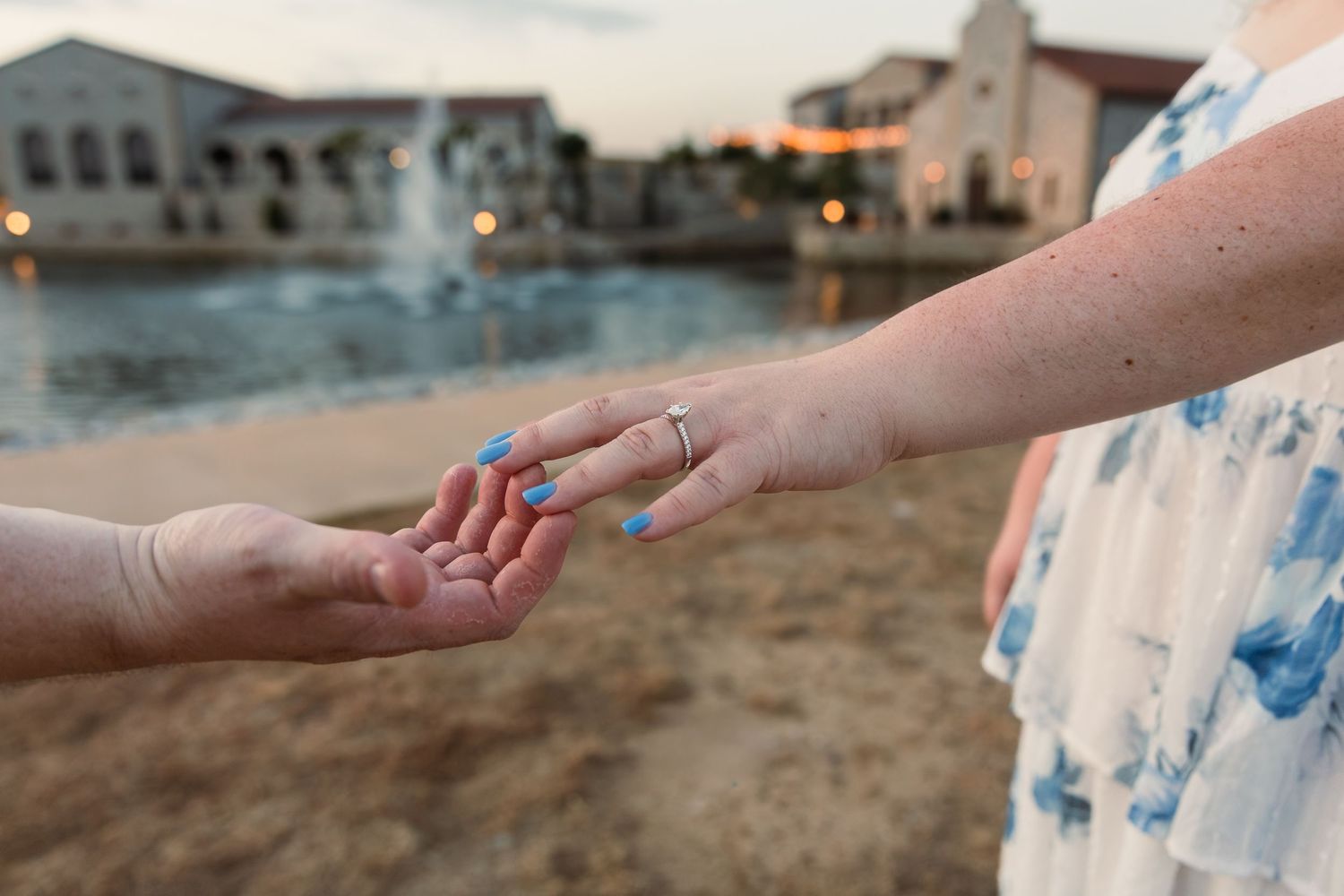 Two women holding hands outdoors near a fountain, one wearing an engagement ring, celebrating a proposal at dusk.