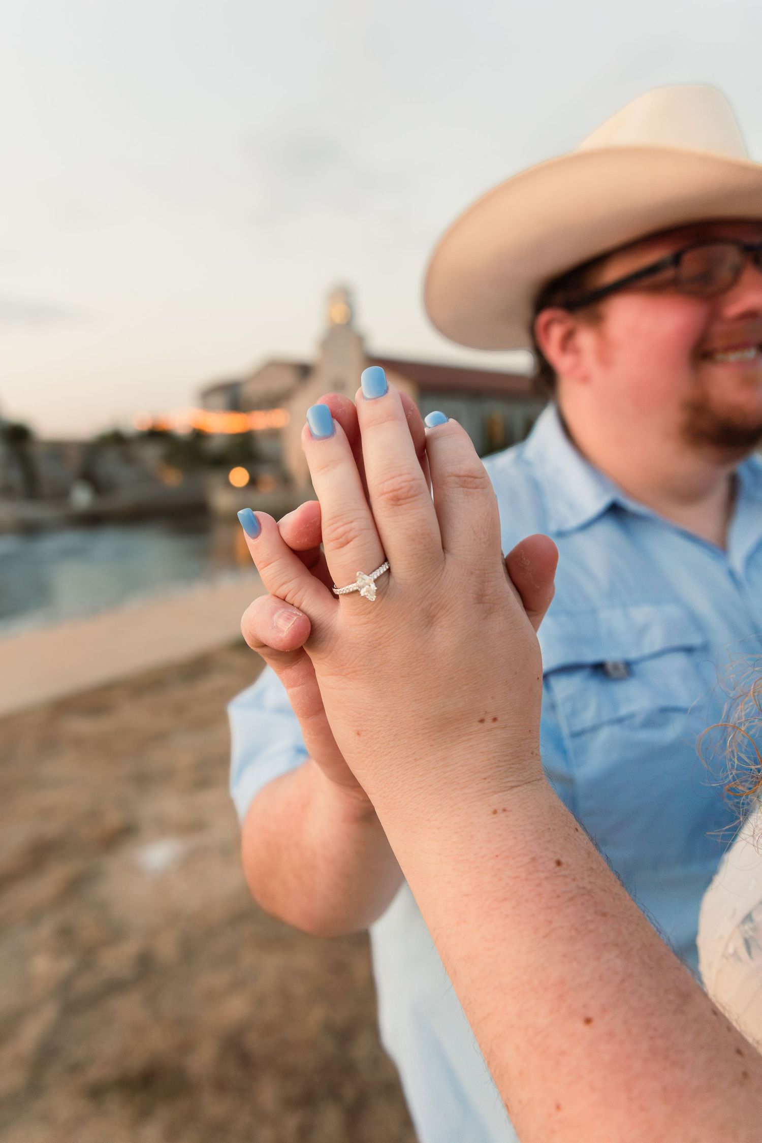 Couple holding hands showing engagement ring with blue nails, man in cowboy hat and blue shirt at sunset beach.
