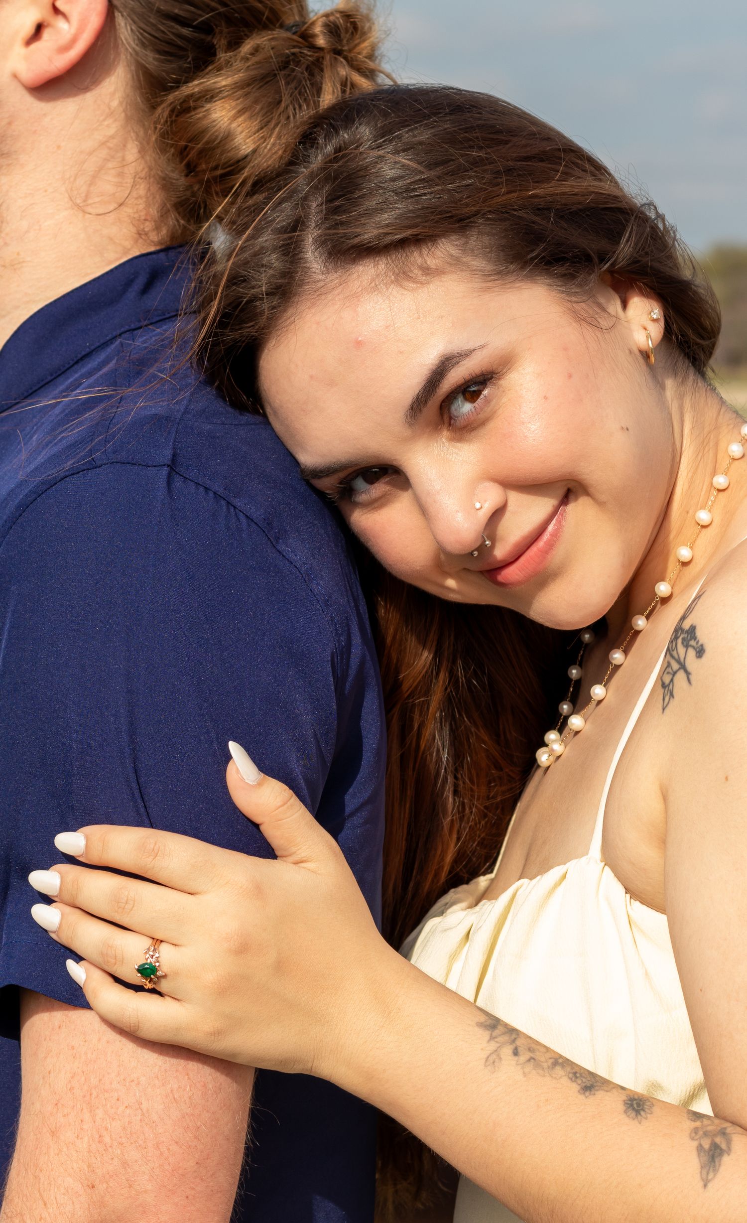 Woman in white dress hugging man, smiling, wearing emerald ring and pearl necklace with tattoo visible.