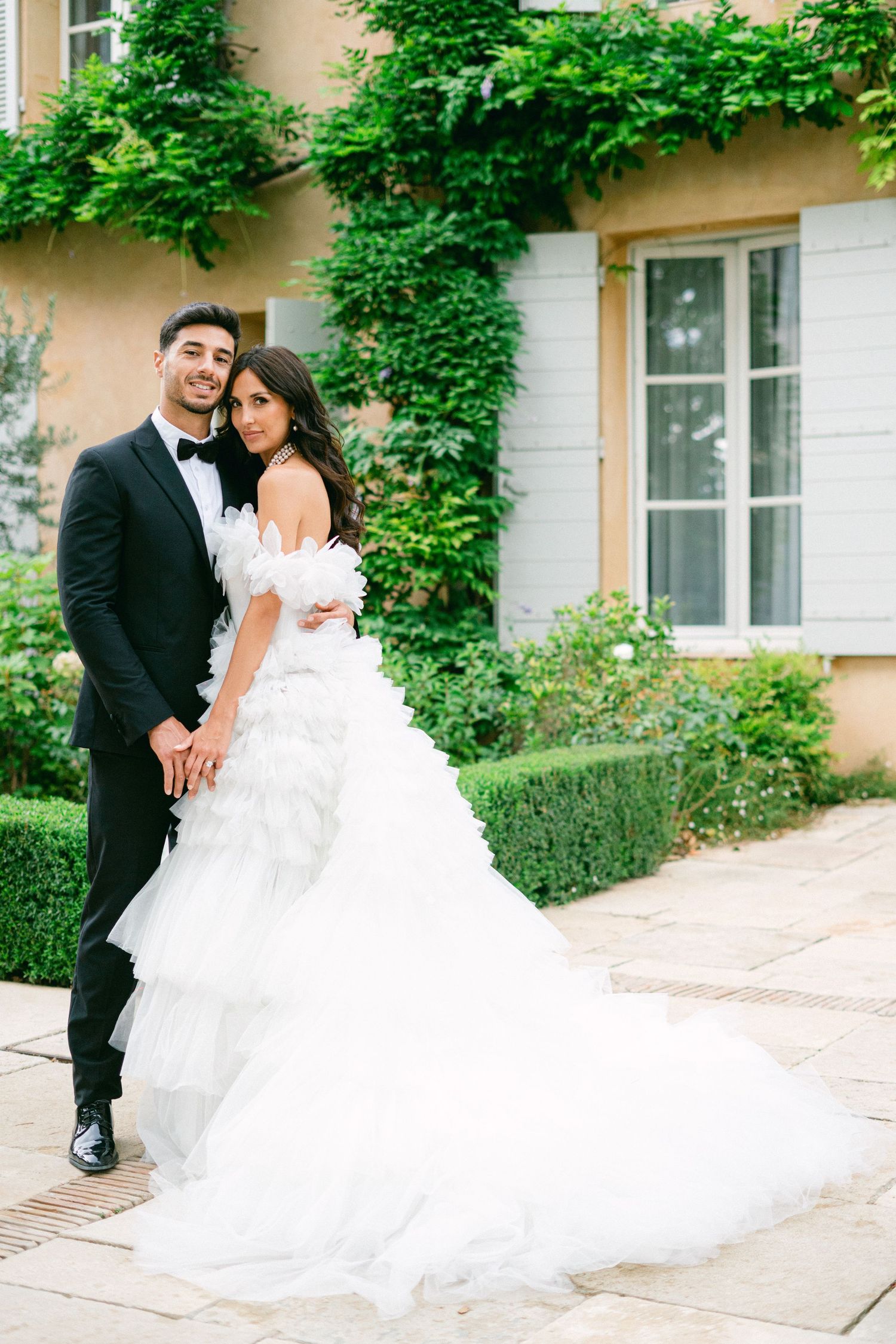 Bride in white ruffled gown and groom in black tuxedo pose together outside an ivy-covered French villa.