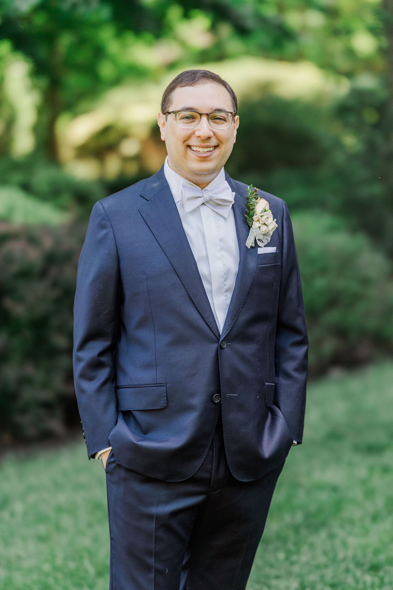 Smiling groom in navy suit with bow tie and boutonniere posing outdoors in lush green garden setting.