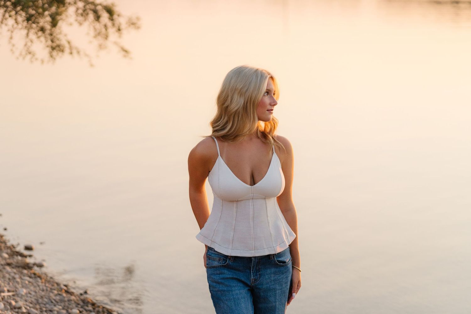 Blonde woman in white tank top and jeans standing by a lake at golden hour sunset.