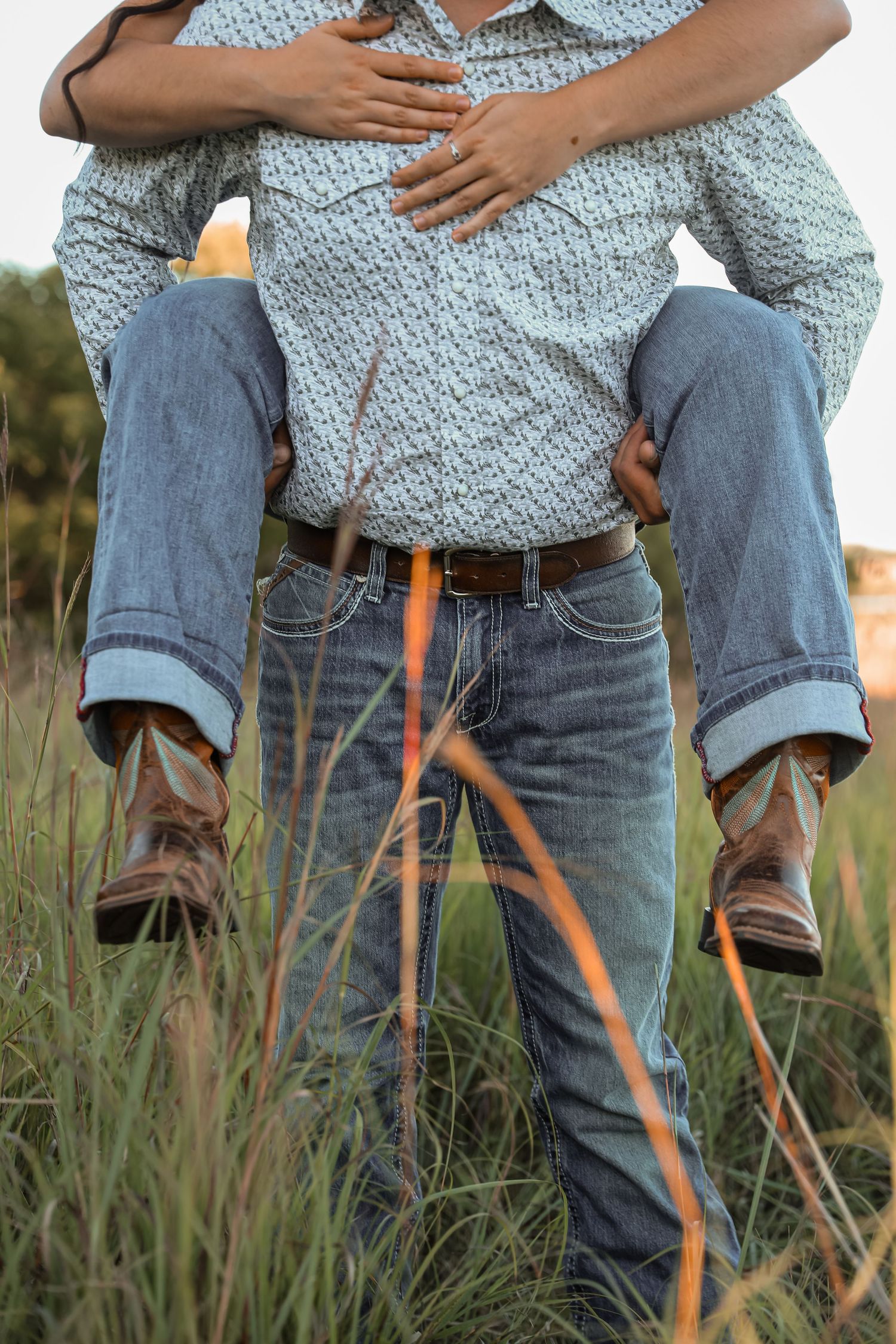 Couple in jeans and cowboy boots sharing a piggyback ride in a grassy field at golden hour.