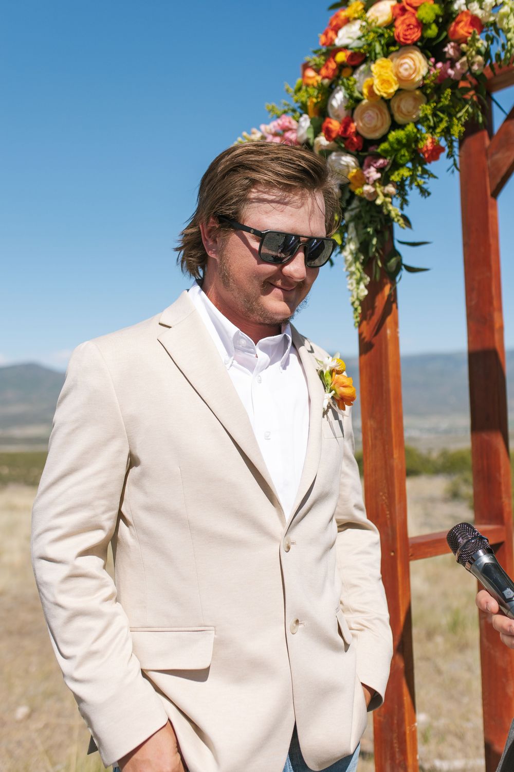 Groom in beige suit with orange boutonniere and sunglasses standing at colorful floral wedding arch outdoors.
