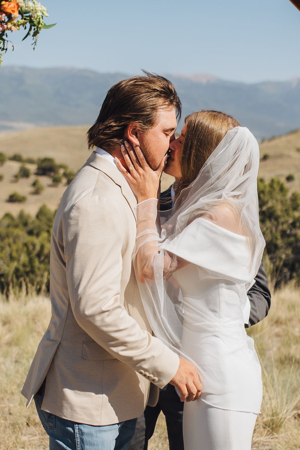 Bride and groom share first kiss at outdoor wedding ceremony with mountain views behind them.