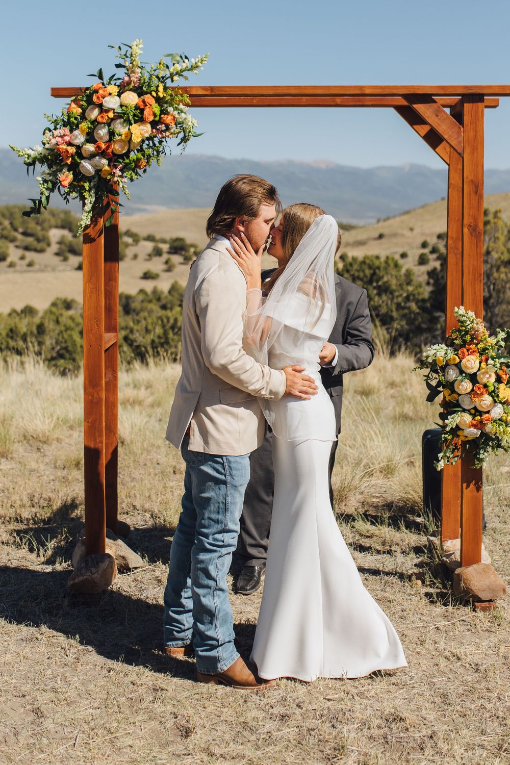 Newlyweds kiss under floral-decorated wooden arch at outdoor wedding with mountain scenery backdrop.